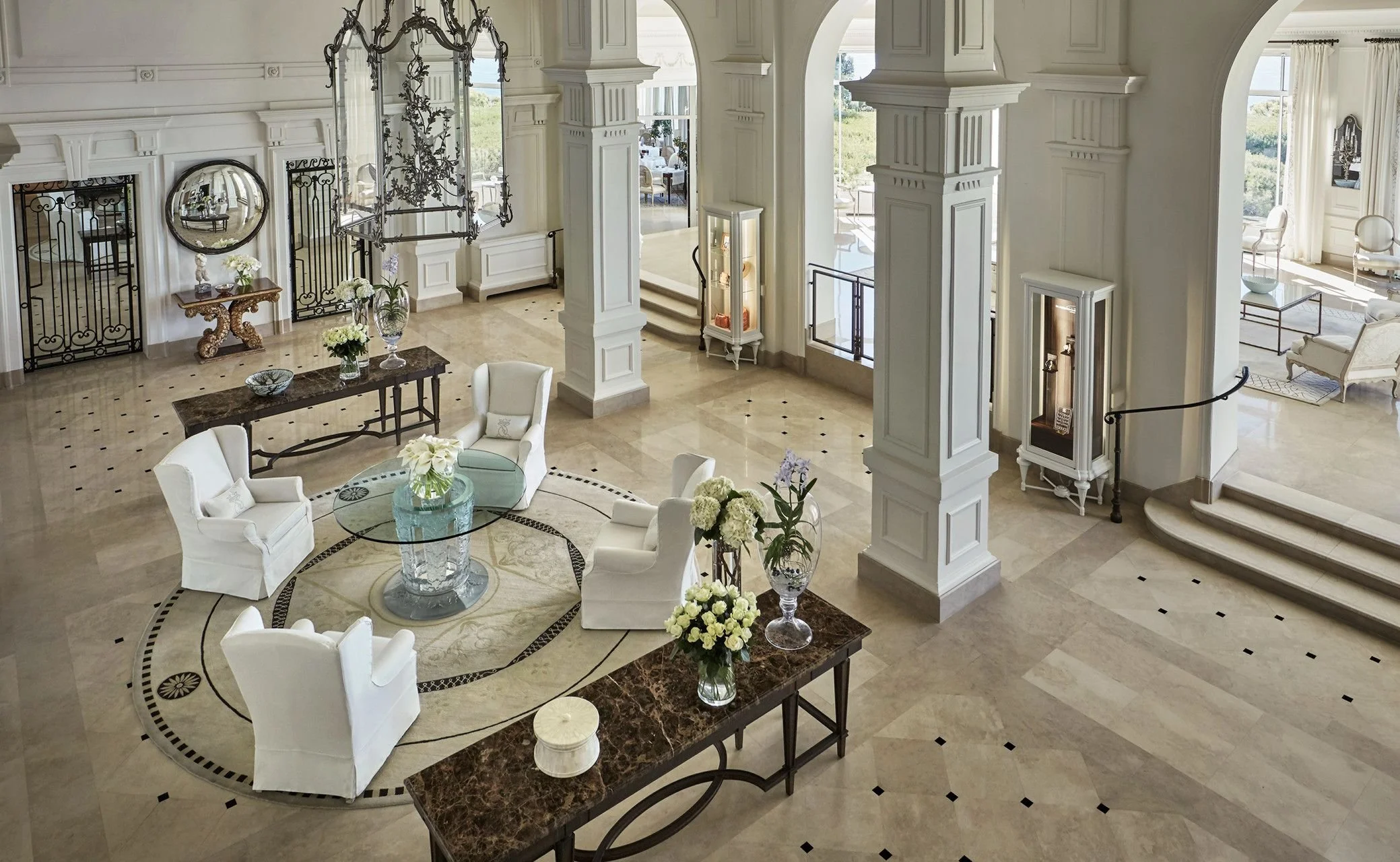 Elegant hotel lobby with white decor, marble floors, and multiple seating areas. Contains columns, large windows with curtains, decorative vases, and tables with flowers.