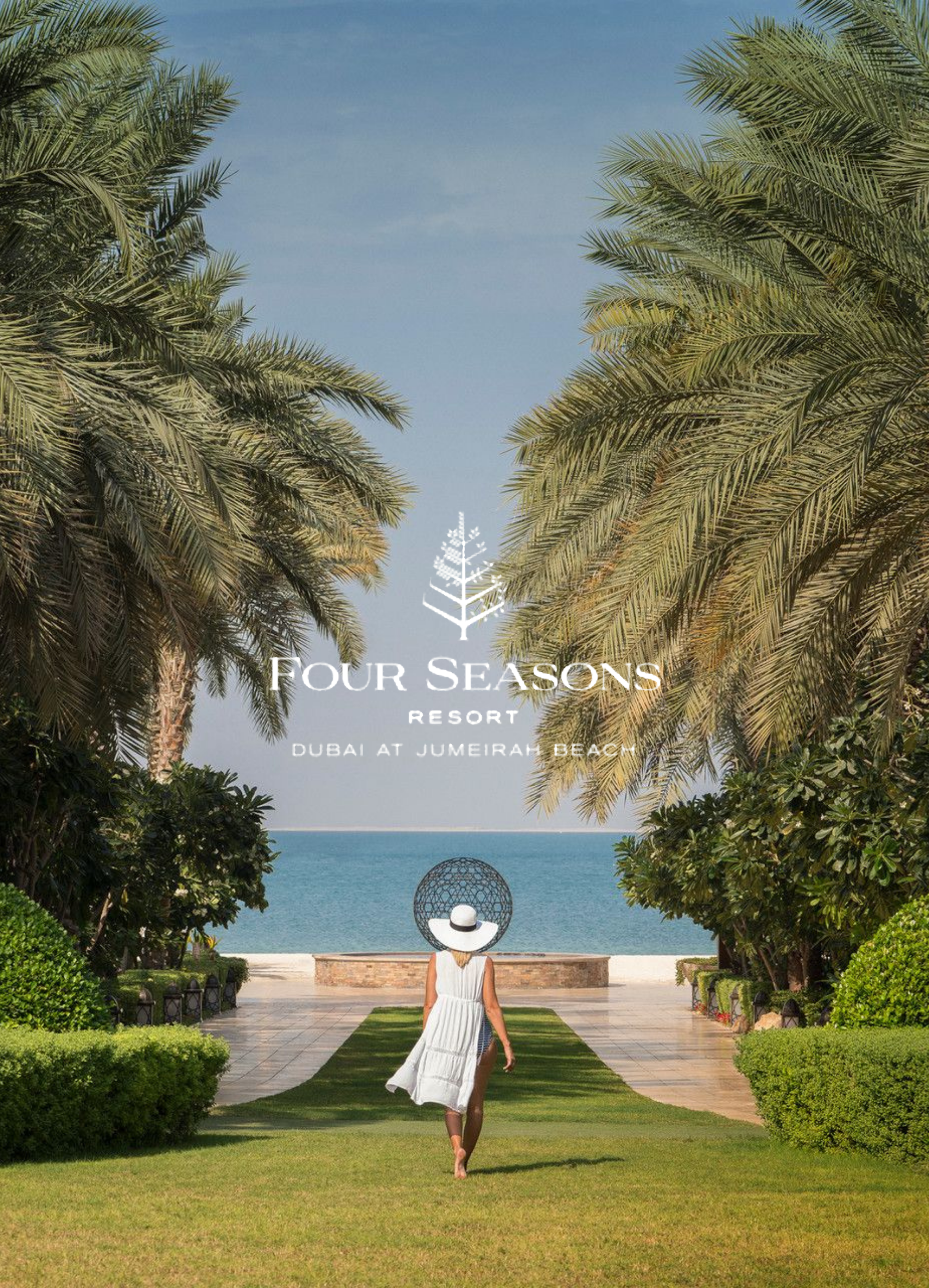A woman in a white dress walking barefoot on a green lawn towards a resort overlooking the ocean, with palm trees on either side and a woman wearing a large sun hat sitting on a bench by the water. The sky is clear and blue.
