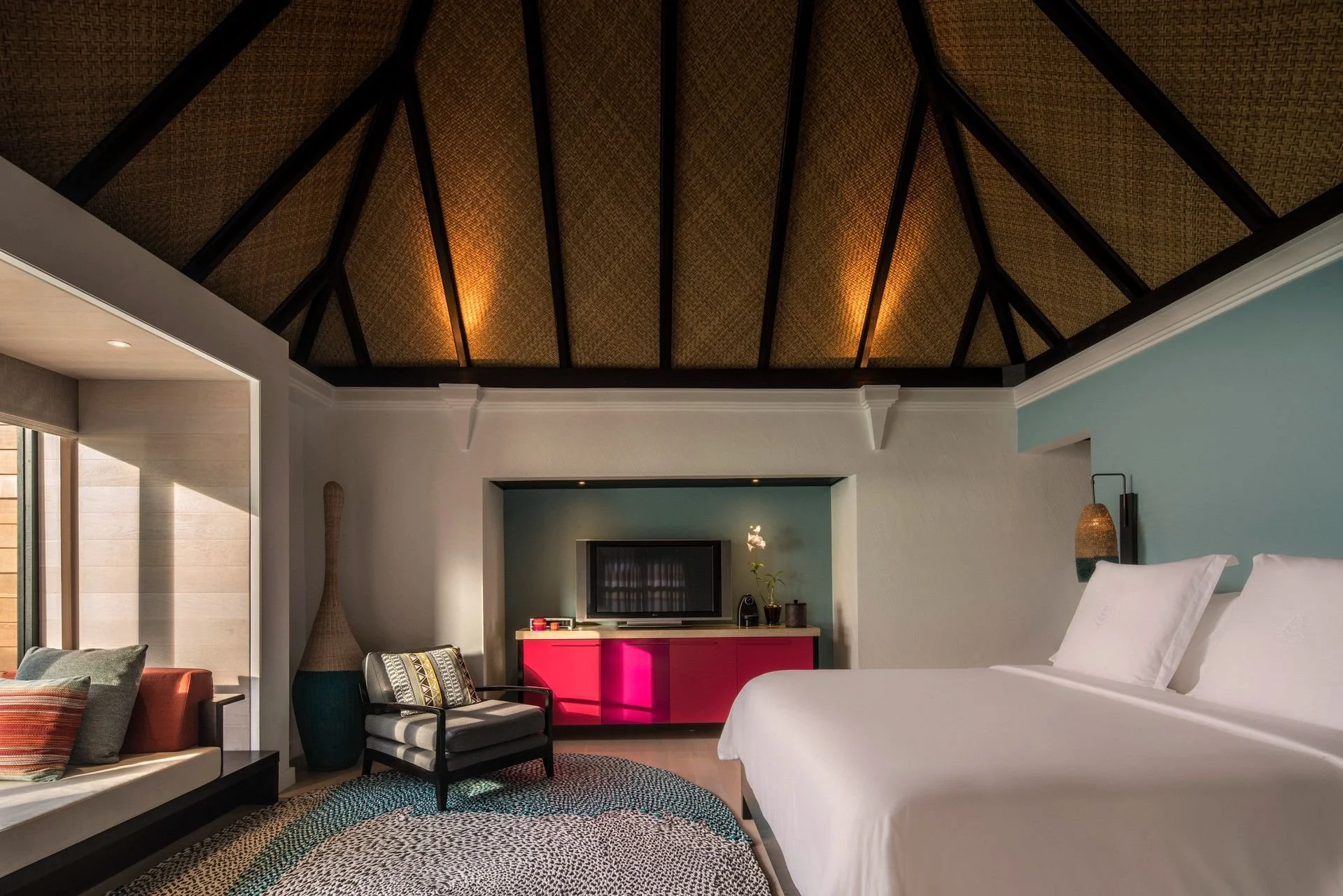 Interior of a modern hotel room featuring a vaulted ceiling with wooden beams, a bed with white linens, a colorful pink cabinet, a television on a wall alcove, a small balcony area with sliding glass doors, and cozy seating with pillows and a woven rug.
