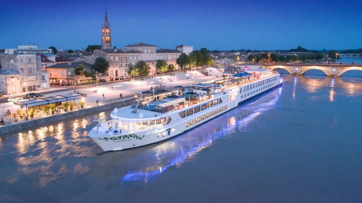 A large white cruise ship sailing along a river at dusk with a illuminated cityscape and historic buildings in the background.