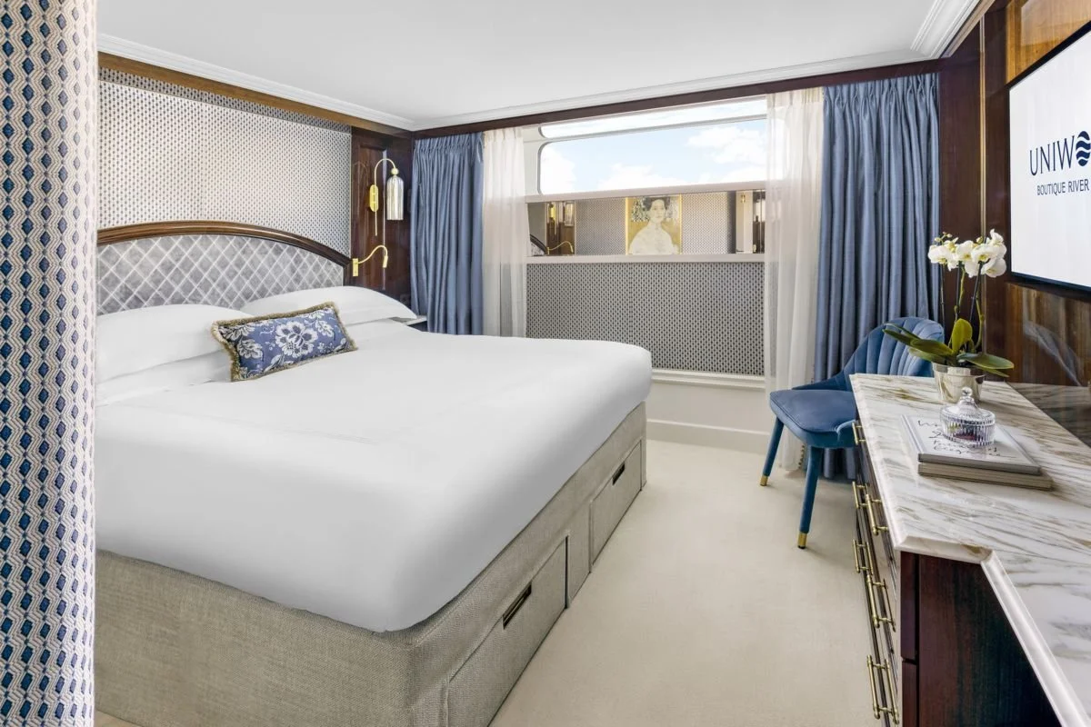 A neatly made hotel room with a large bed, blue and white decor, a window with curtains, a marble-topped desk, a chair, and a flat-screen TV on the wall.