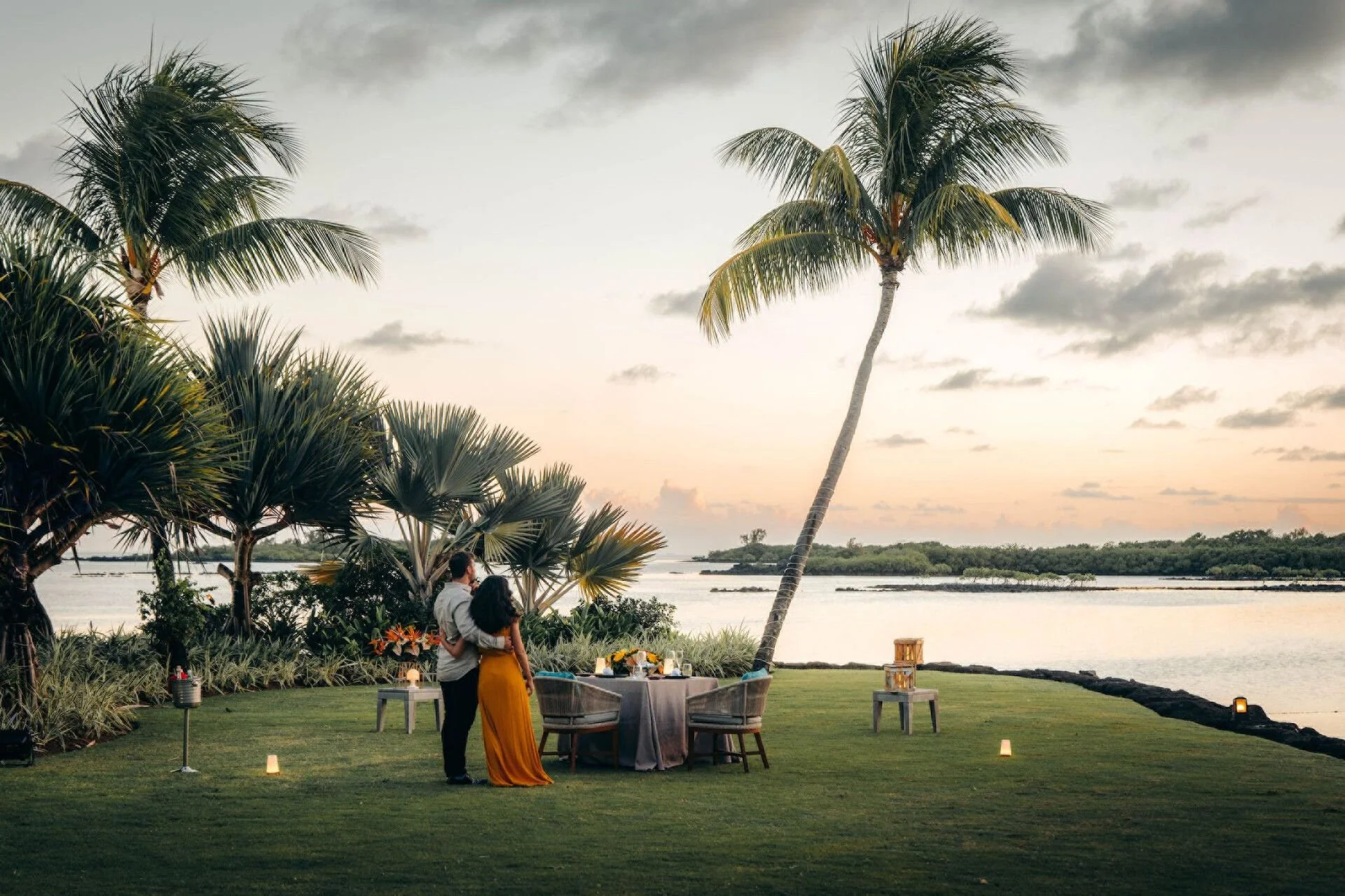 A couple stands close together near a table set for a romantic dinner outdoors, with palm trees and a river in the background during sunset.