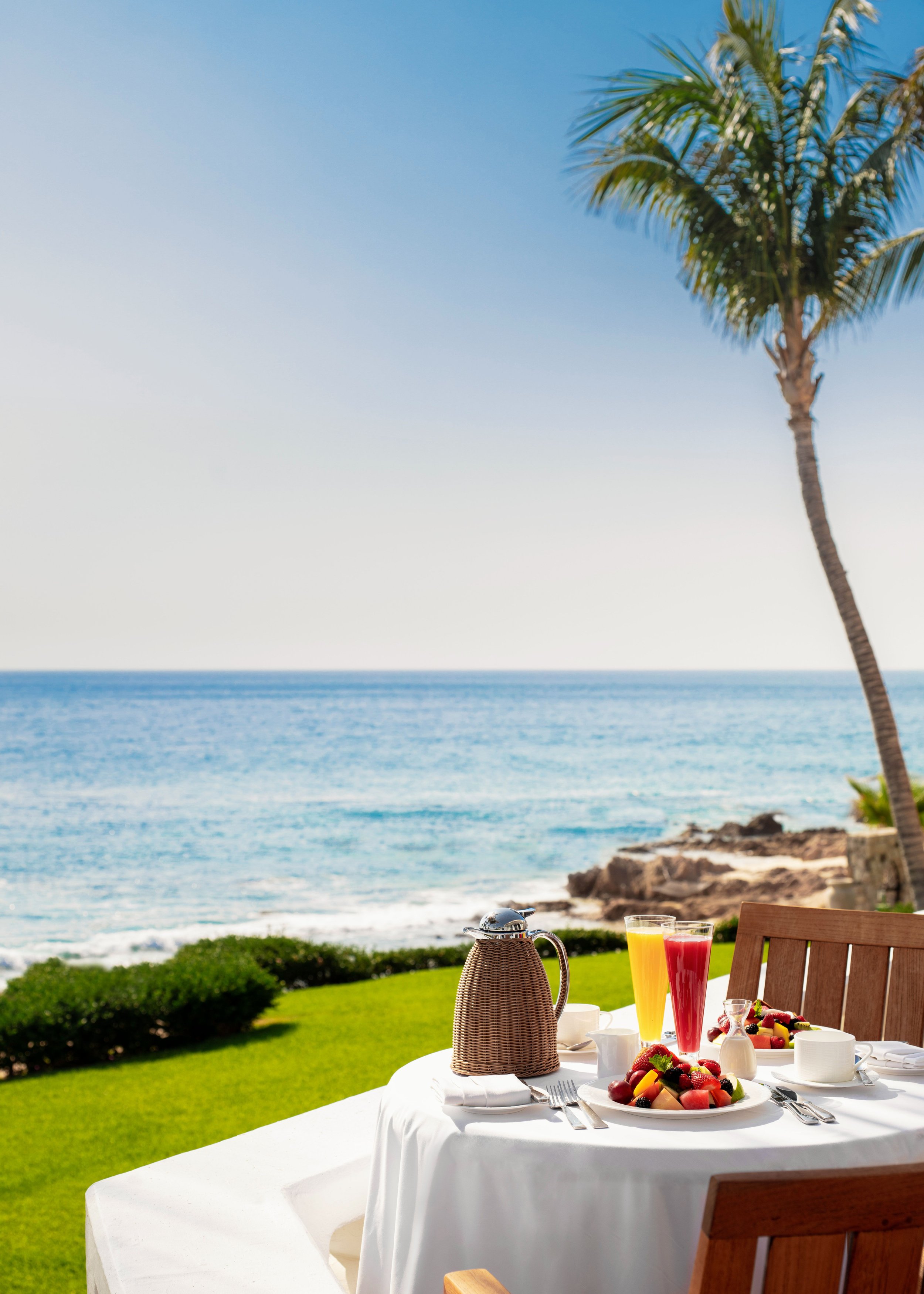A table set for breakfast outdoors by the ocean, with a white tablecloth, fruit platter, glasses of juice, and woven basket, with a palm tree and ocean view in the background.