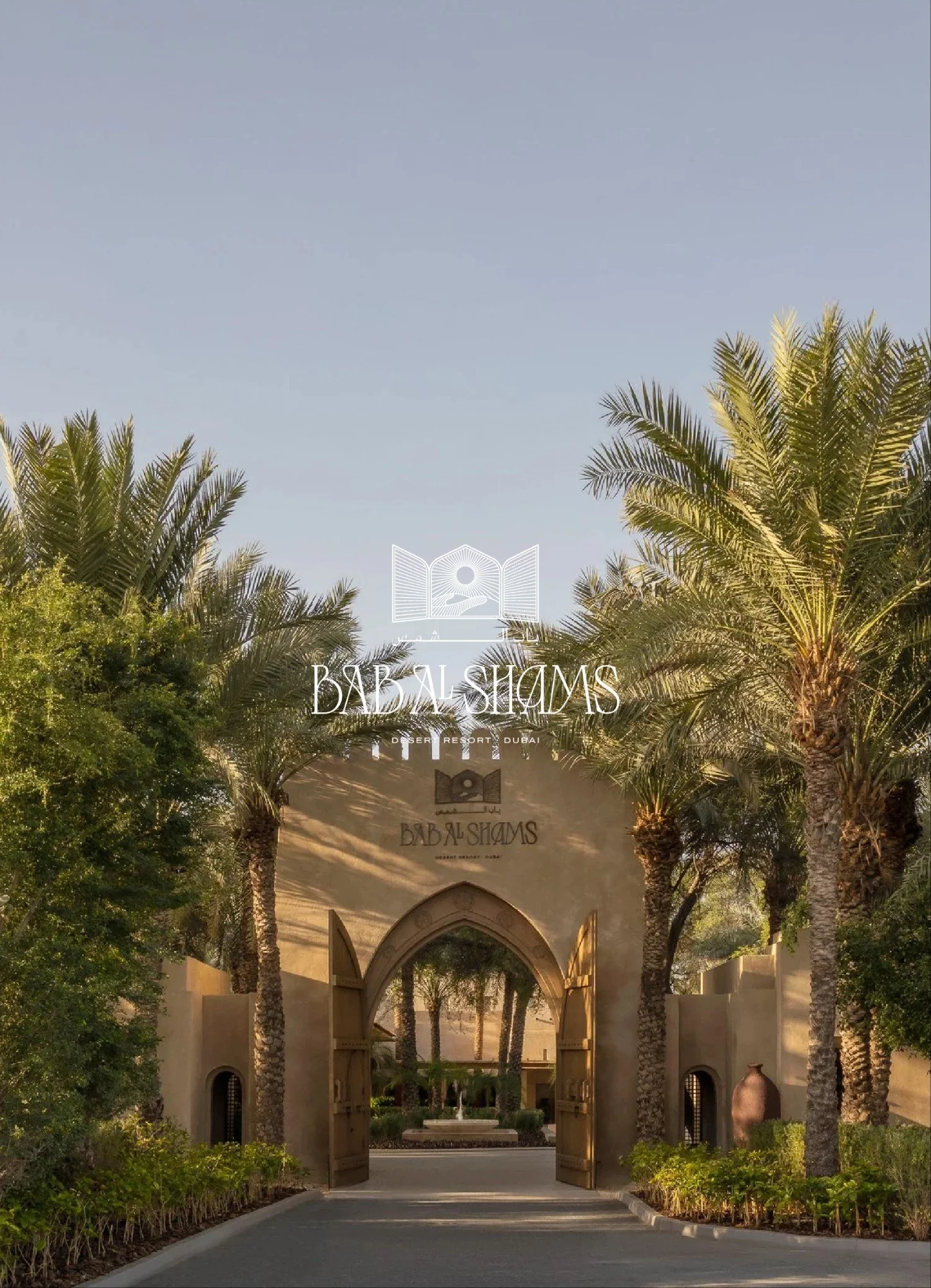 Entrance to Bab Al Shams Desert Resort in Dubai, with an ornate archway surrounded by tall palm trees and lush greenery, under a clear blue sky.