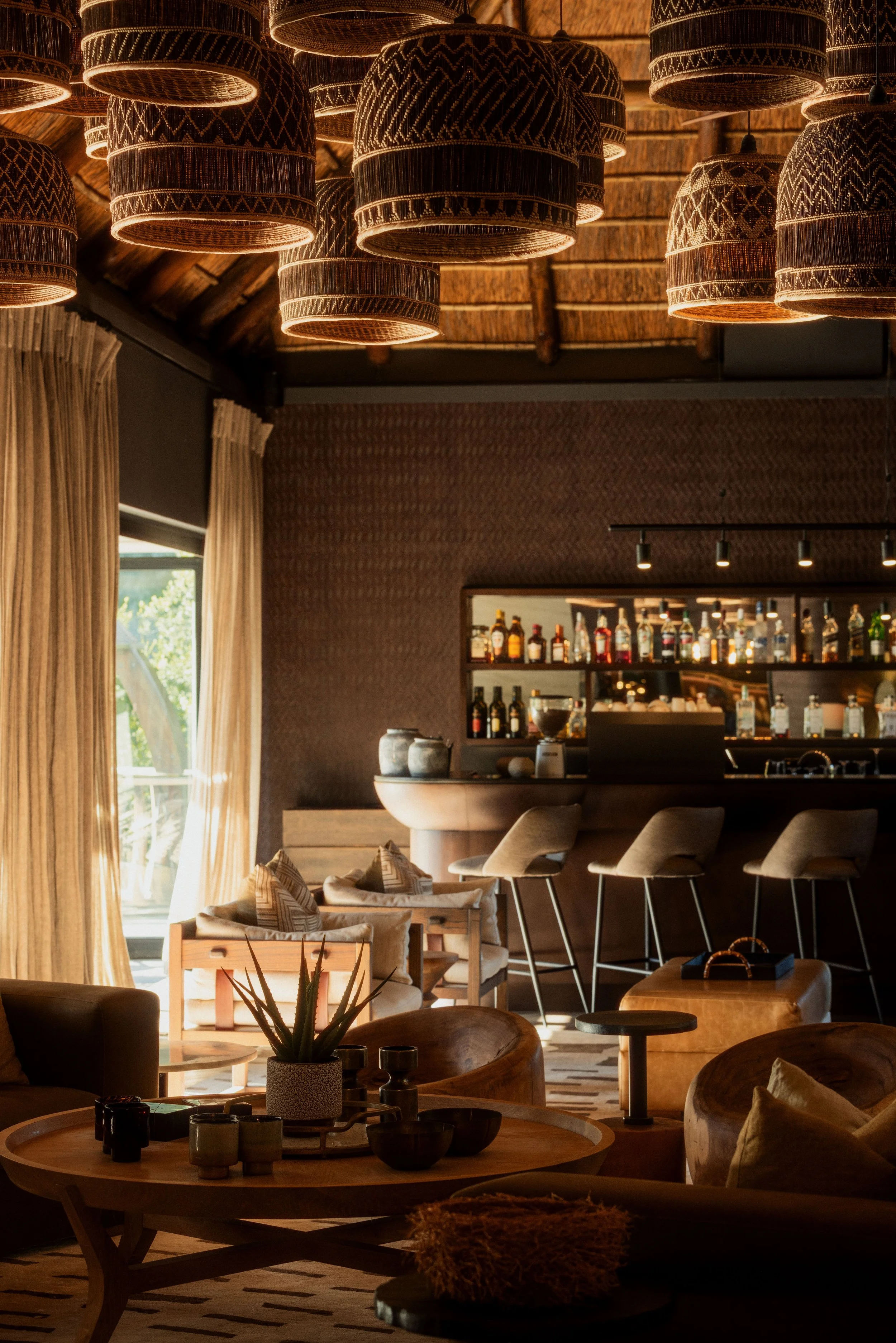 Interior of a cozy bar or lounge with warm lighting, wooden furniture, a bar counter with liquor bottles, and decorative woven lampshades hanging from the ceiling.