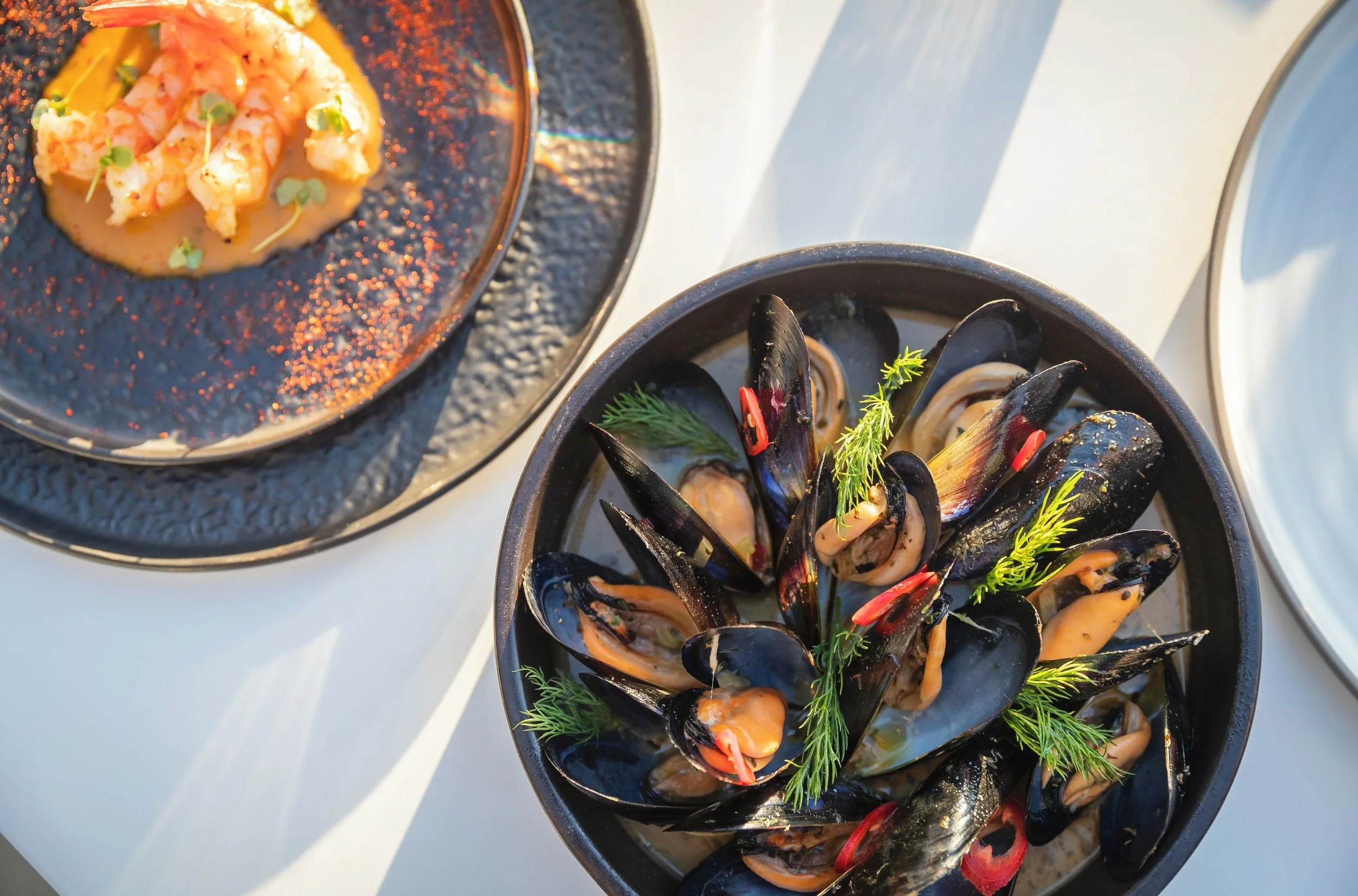 A close-up of three black bowls containing seafood dishes on a white surface. The top bowl has cooked shrimp garnished with microgreens and served with a sauce. The middle bowl contains cooked mussels, garnished with herbs and thin slices of red chili.