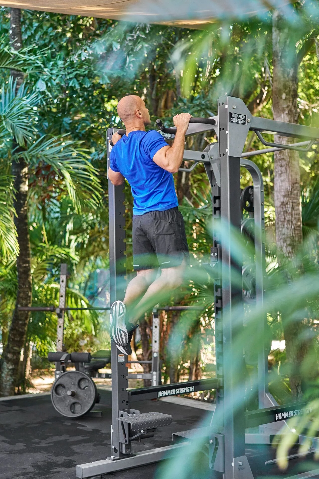 A man in a blue shirt and black shorts performs a pull-up on a gym station outdoors surrounded by green plants and trees.