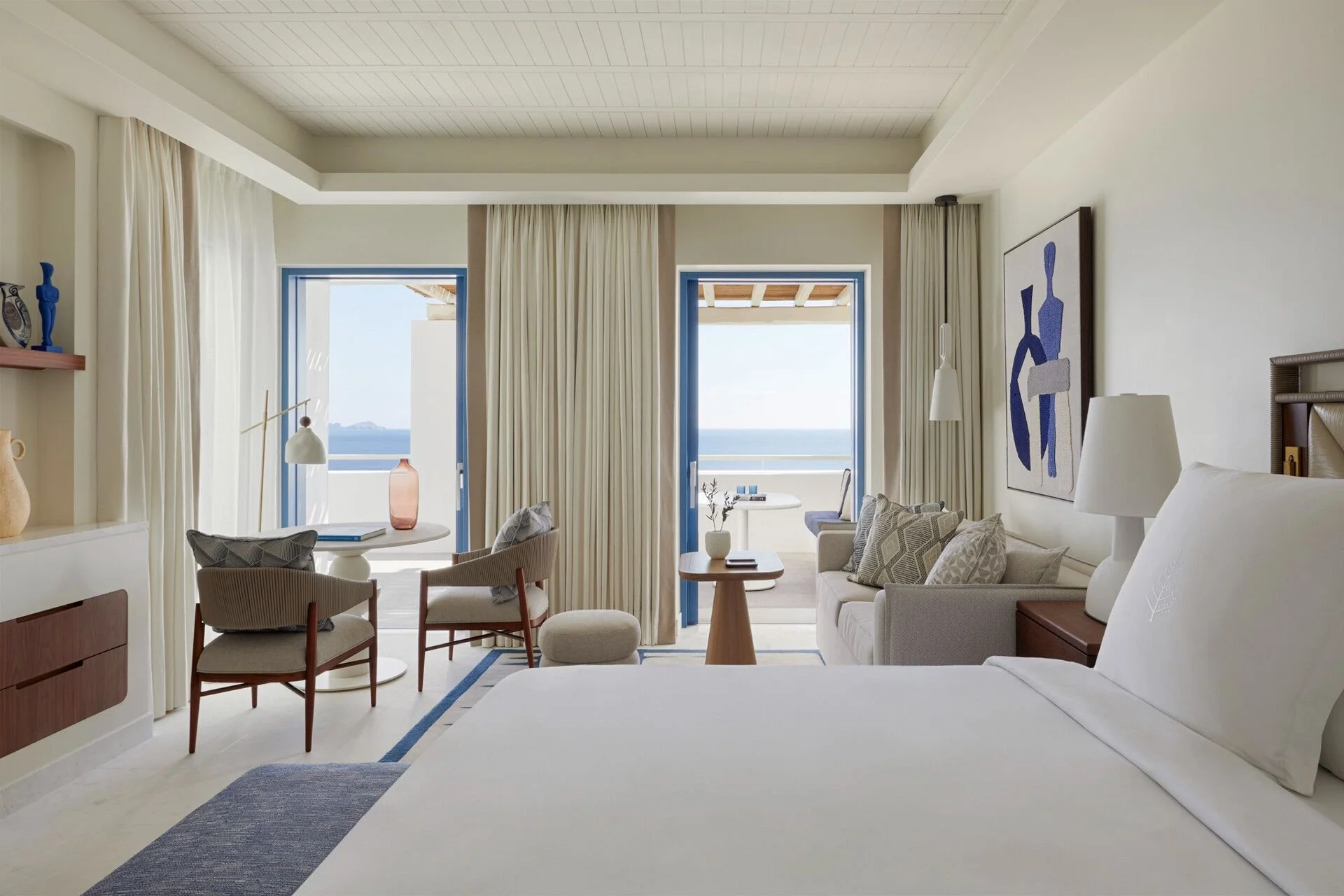 A bright hotel room with ocean views through two open balcony doors, furnished with a white bed with decorative pillows, a sitting area with chairs and a small table, and modern art on the walls.