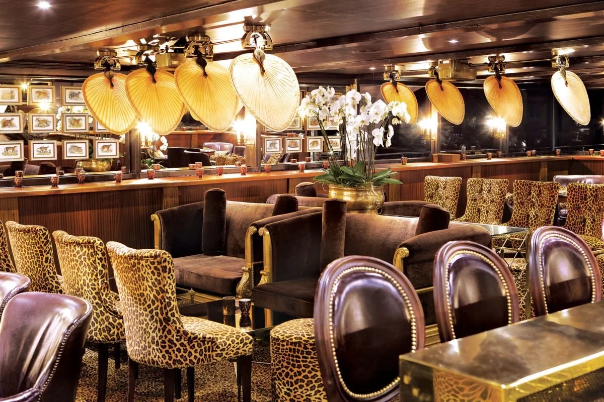 Luxurious restaurant or lounge with leopard print chairs and velvet sofas, large white orchids centerpiece, framed animal artwork on dark wood walls, and umbrella-shaped light fixtures hanging from the ceiling.
