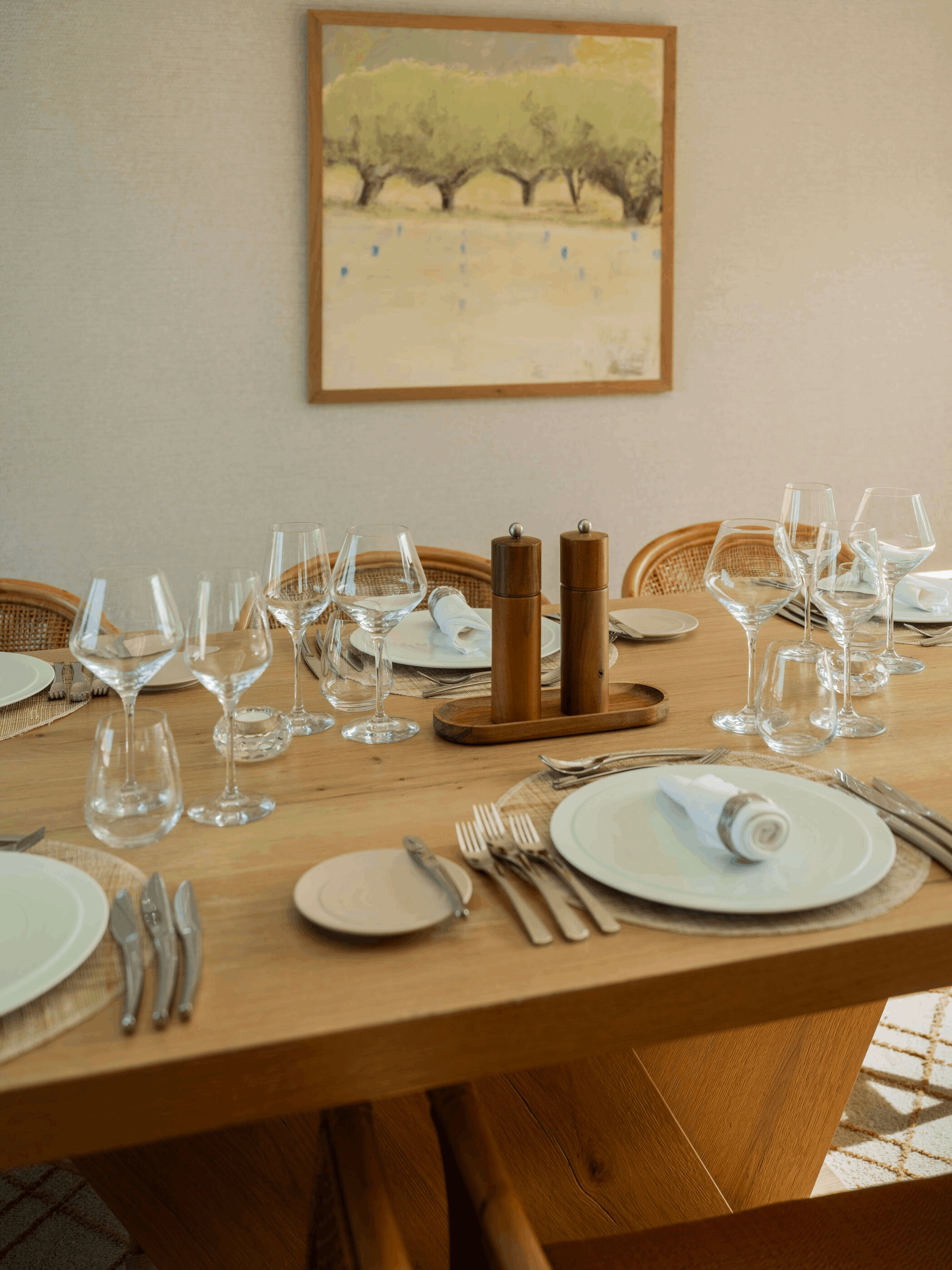A dining table set with plates, silverware, wine glasses, napkins, and salt and pepper shakers, in a room with a framed picture of trees hanging on the wall.