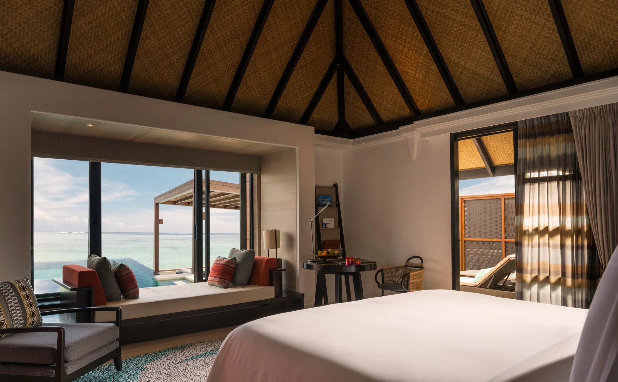 Interior of a tropical beachfront hotel room with large windows, showing a balcony and ocean view, with seating area, chairs, and curtains.