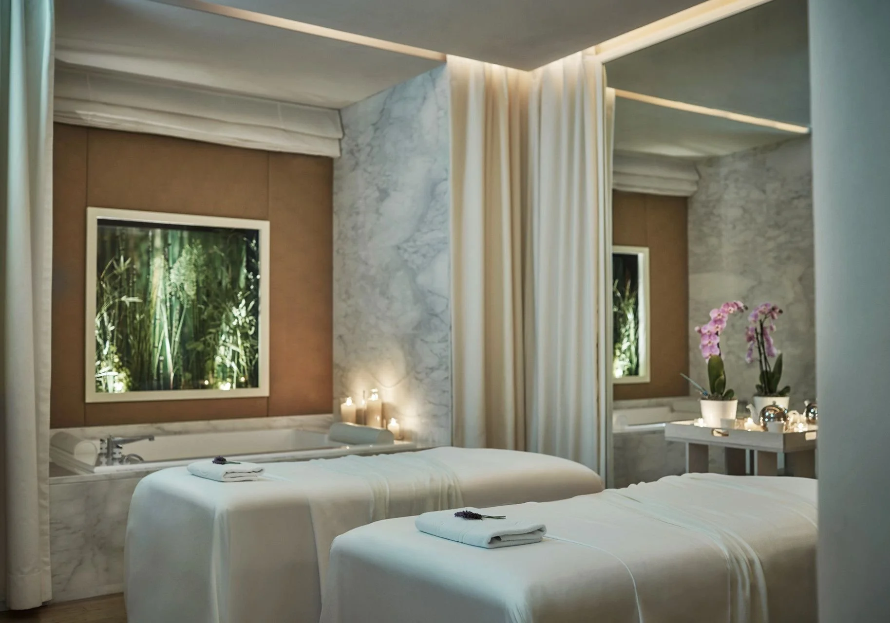 A luxury spa or massage room with two massage beds covered with white sheets and towels, set against marble walls with tropical plant images, pink orchids on a low table, and lit candles creating a relaxing atmosphere.