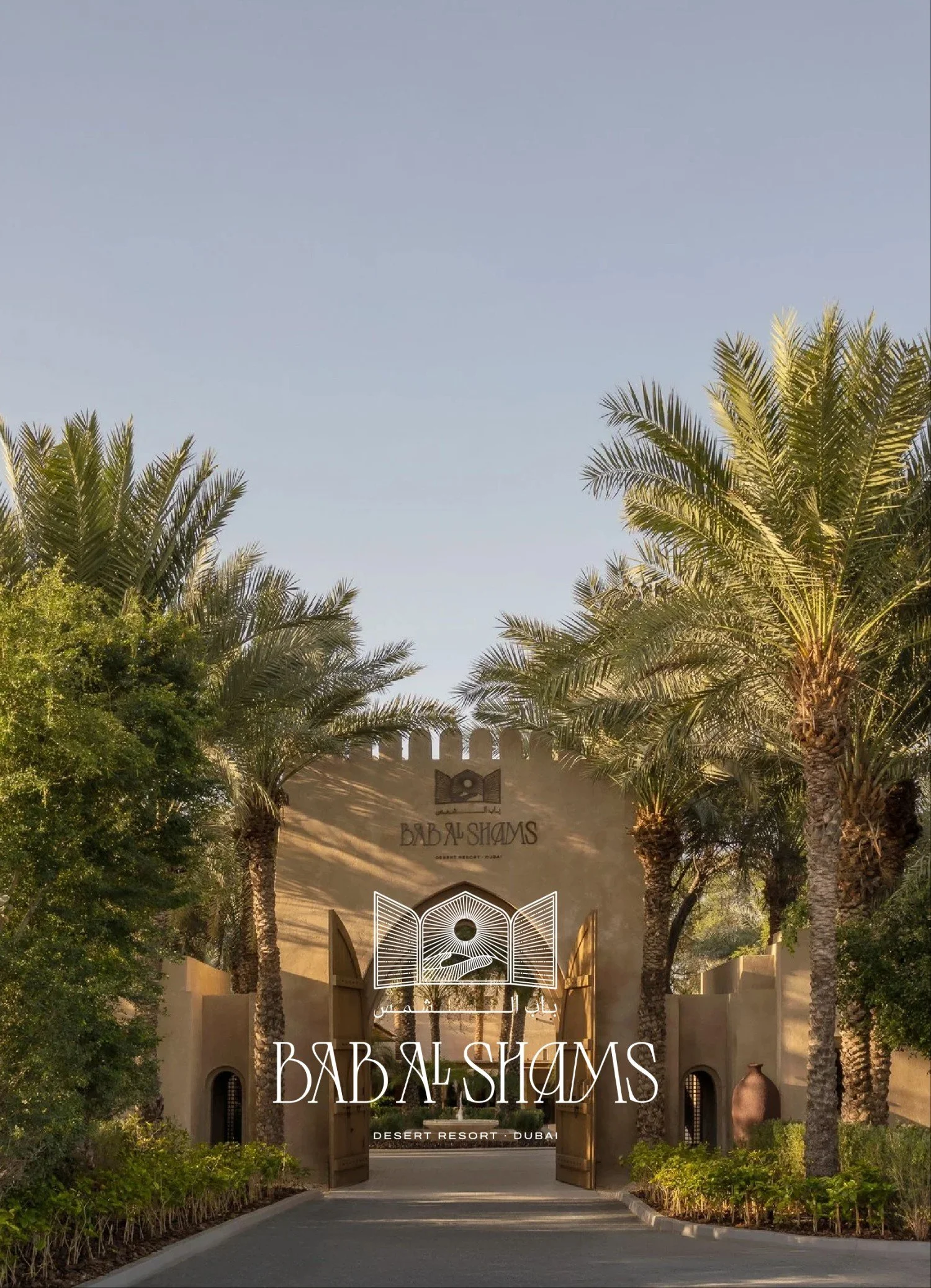Entrance gate to Bab Al Shams Desert Resort Dubai, surrounded by palm trees and lush greenery, with clear sky overhead.