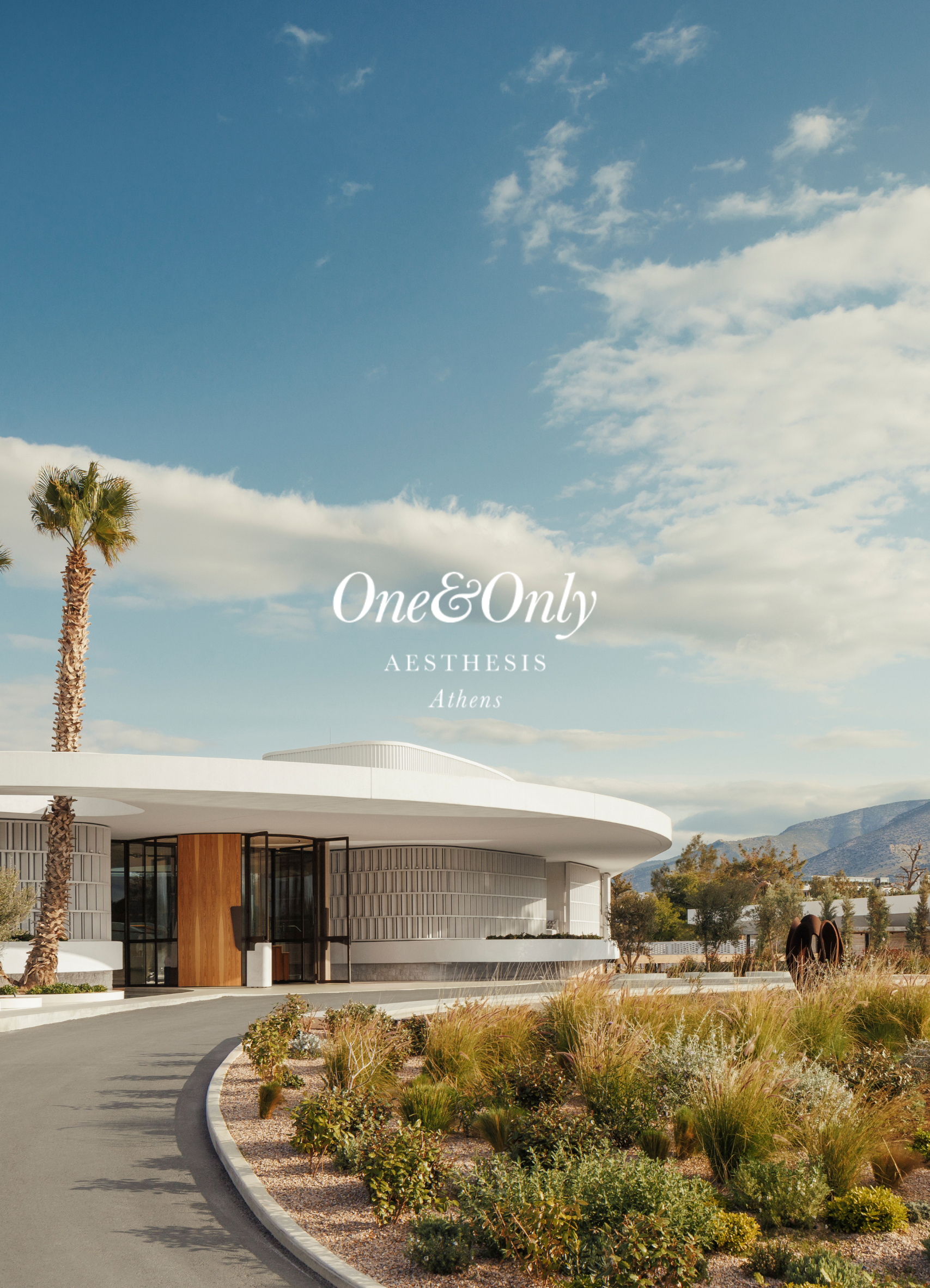Modern architectural building with curved white roof, tan wooden accents, glass walls, and landscaped desert plants in the foreground under a blue sky with clouds, with mountains in the background.