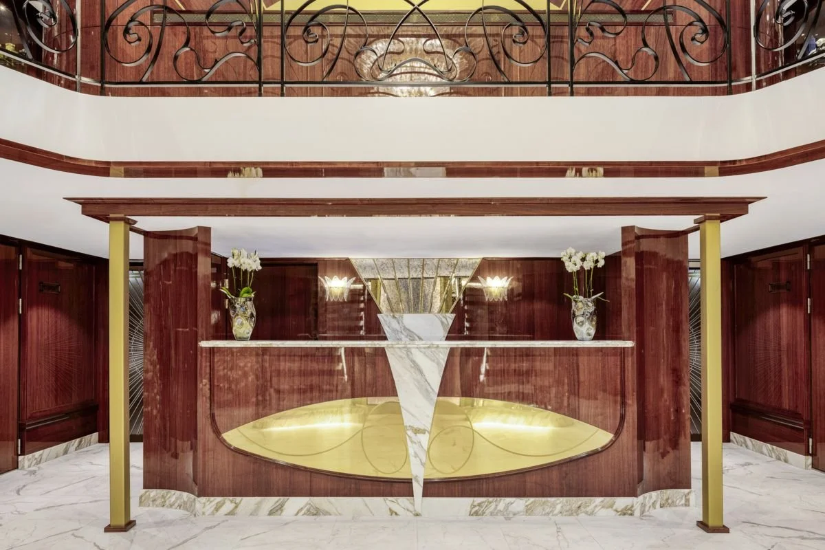 Elegant hotel or building lobby reception desk with polished wooden surfaces, marble accents, and decorative vases with flowers on marble countertop, decorative lighting fixtures, and a wrought iron balcony seen at the top.