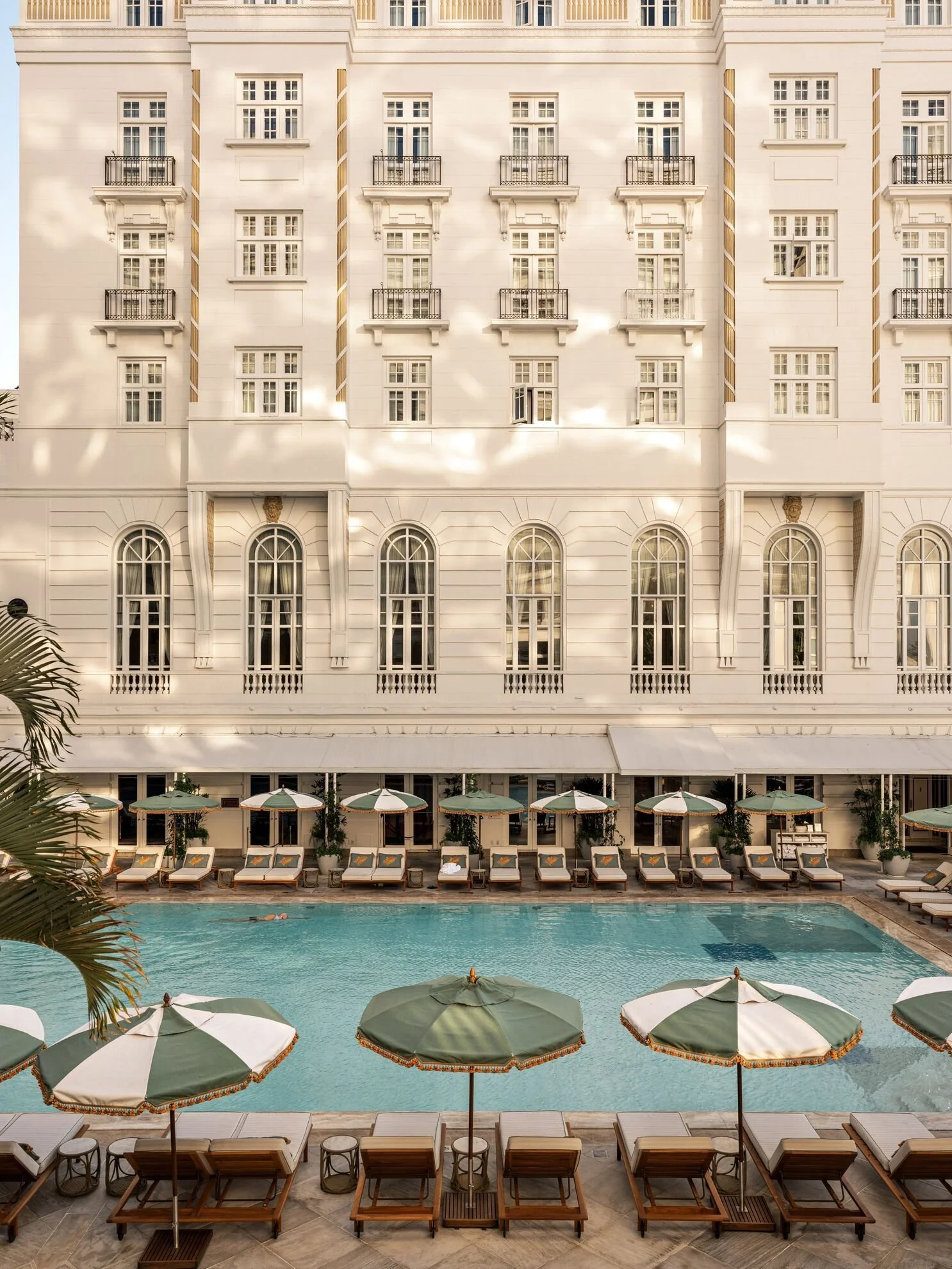 A view of a hotel pool area with sun loungers and umbrellas, with a large elegant building in the background.