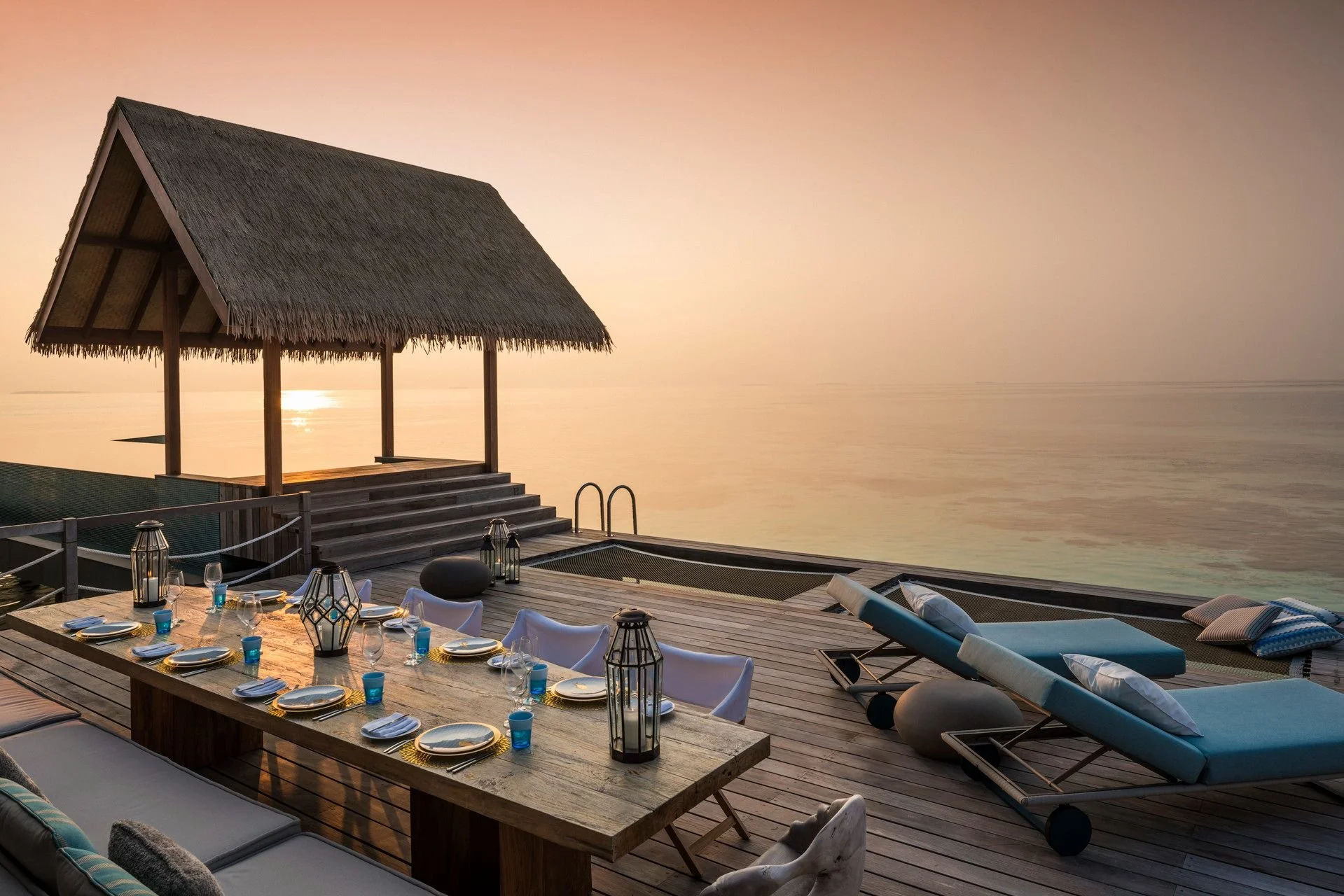 Outdoor dining area on a wooden deck with a long table set with plates, glasses, and lanterns. There are lounge chairs and a thatched-roof hut overlooking a calm body of water during sunset.
