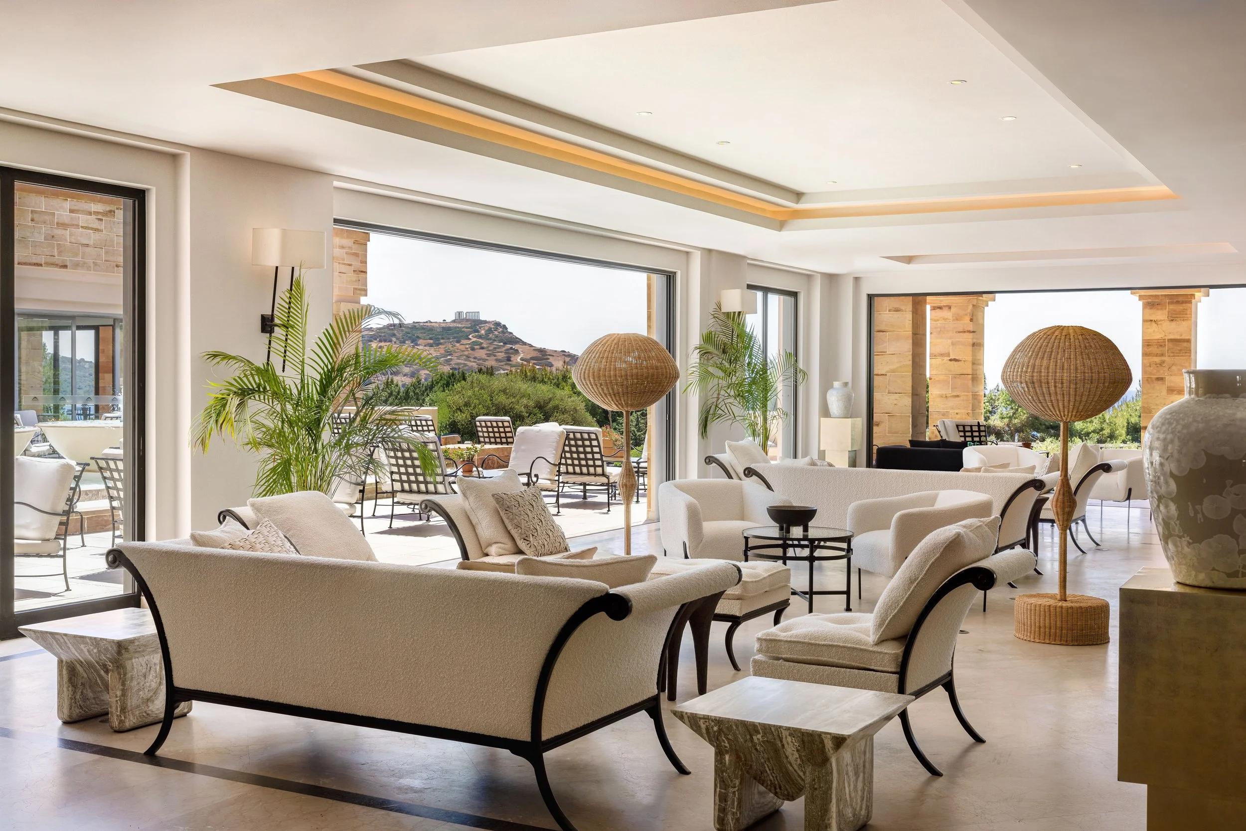 Living room with white sofas, armchairs, and decorative lamps, featuring large windows overlooking an outdoor terrace with chairs and a view of hills and a monument in the distance.