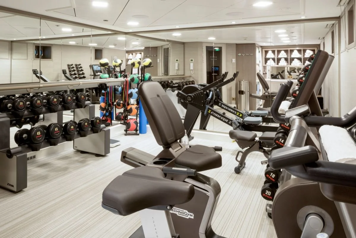 A modern gym with various exercise equipment, including dumbbells, medicine balls, weight machines, and cardio machines, all arranged in a clean, well-lit space with mirror walls.