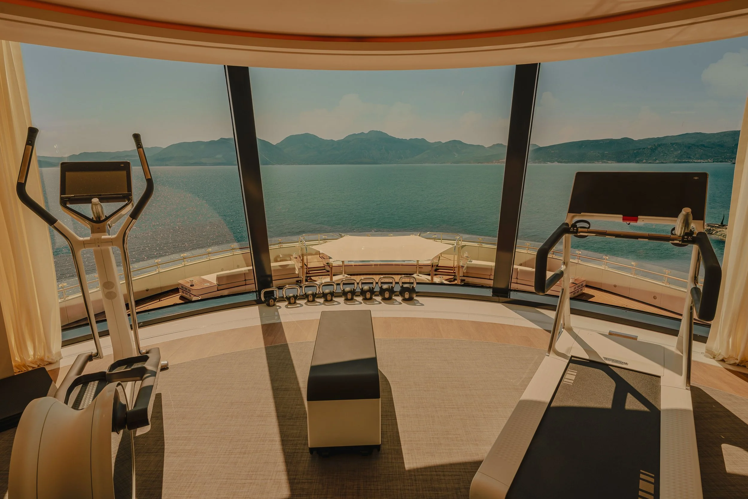 Inside a luxurious cruise ship gym with large windows showing a scenic view of a lake and mountains, exercise equipment includes elliptical machines and a treadmill.