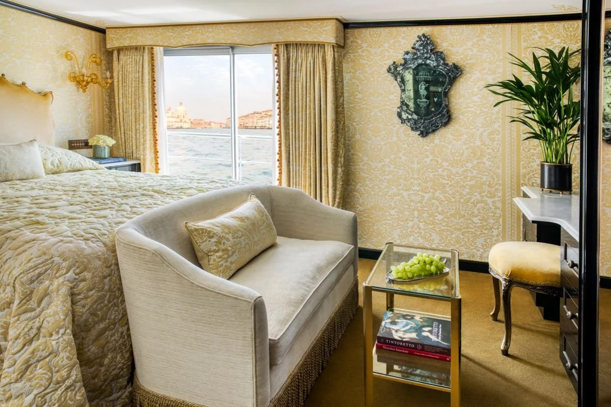 Luxurious hotel room with a large bed, a view of a canal outside the window, and vintage furniture, including a cream-colored sofa, a glass-topped side table with grapes and books, a potted plant, and decorative wall art.