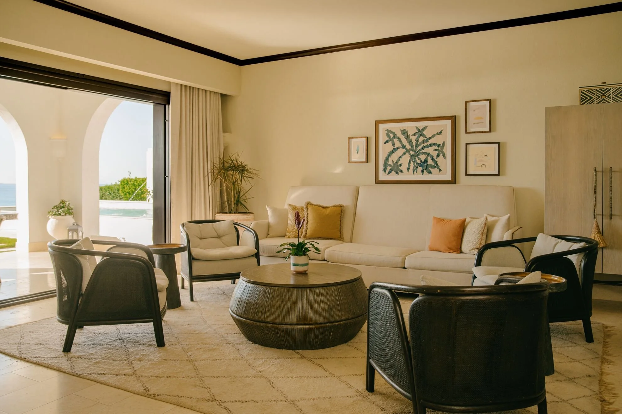 A living room with a large beige sofa decorated with throw pillows, a round coffee table with a potted plant, and multiple framed plant-themed artworks on the cream-colored wall. There are multiple chairs surrounding the coffee table, and a sliding glass door that opens to a balcony with an ocean view and lush greenery.
