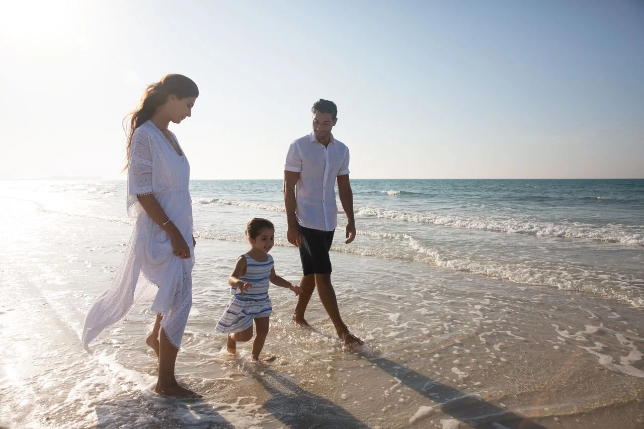 A family of four walking along the shore at the beach, with the ocean and clear sky in the background.