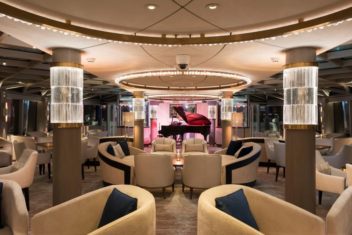 Elegant lounge with armchairs and sofas arranged in a circle, a red grand piano on a stage in the background, and modern lighting fixtures on columns, creating a luxurious atmosphere.