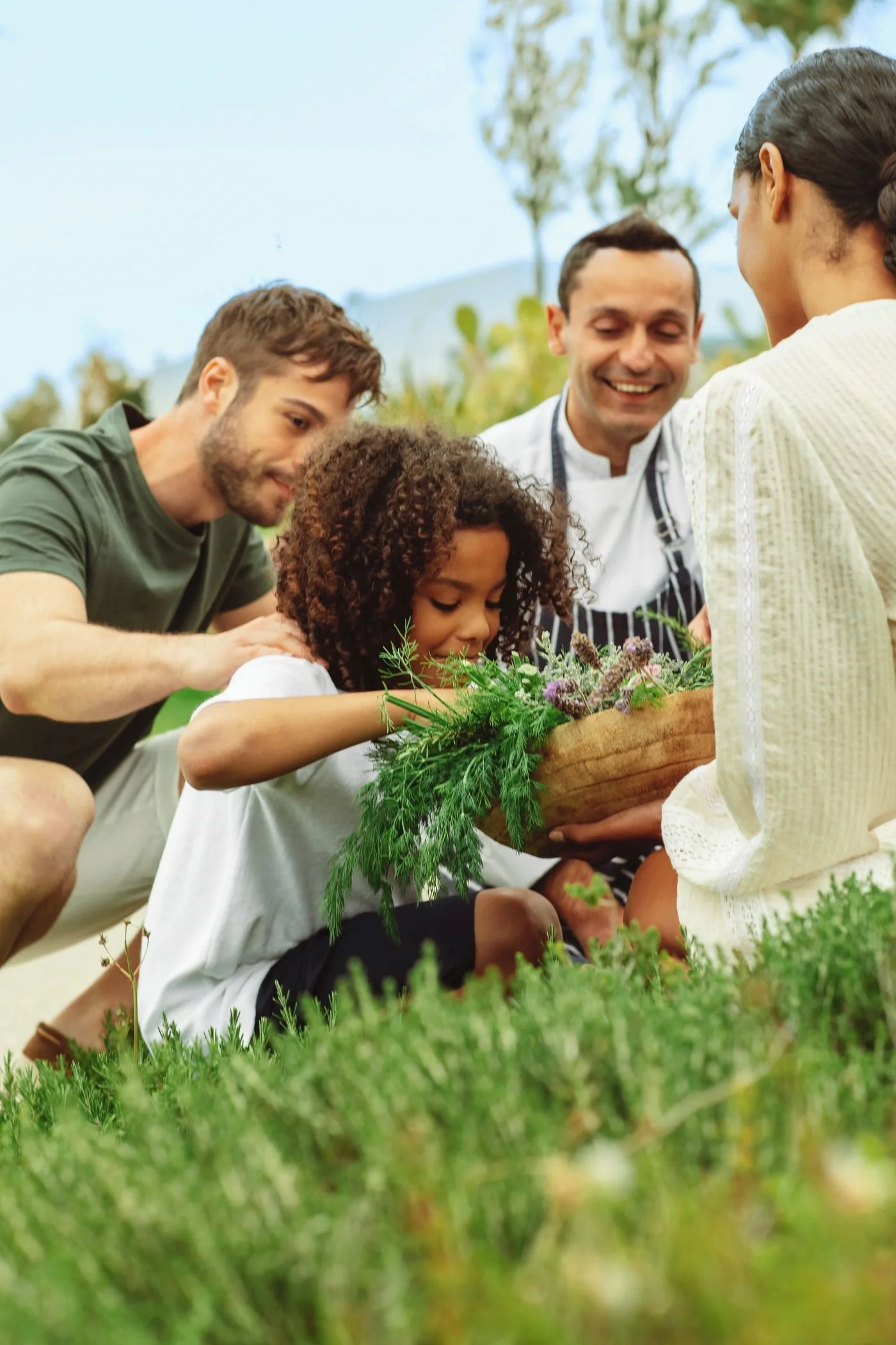 A group of people, including a young girl, are gathered outdoors in a garden, examining a basket of fresh herbs and flowers with a woman. They are smiling and appear to be gardening or participating in a nature activity.