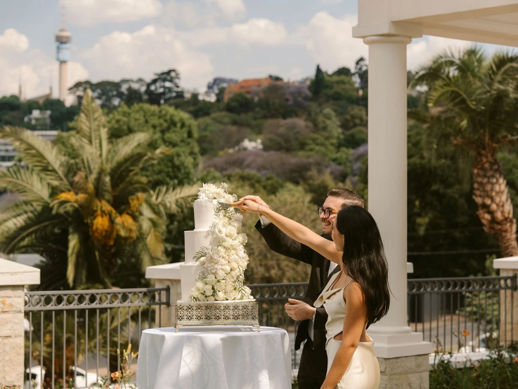 A couple is cutting a wedding cake outdoors on a balcony with trees and a cityscape in the background. The bride and groom are smiling, and the man is wearing glasses and a tuxedo.