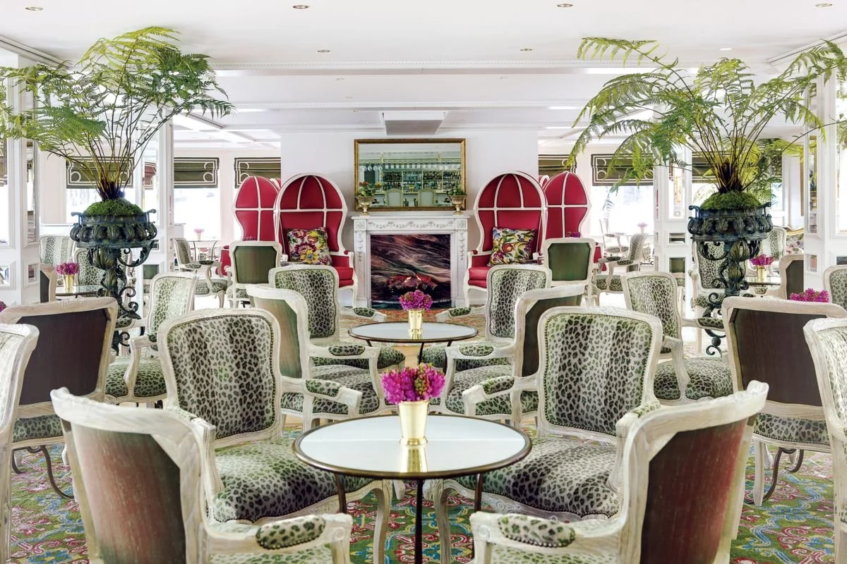 Elegant hotel lounge with leopard print armchairs, round tables with pink floral centerpieces, and large potted ferns.