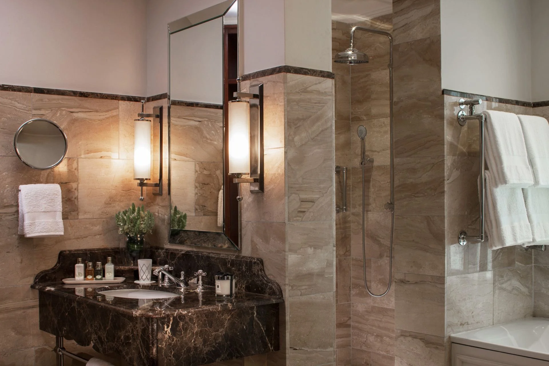 Bathroom with marble tiles, a dark marble countertop vanity with toiletries, a large mirror, wall-mounted light fixtures, a small round mirror, towels on a towel rack, and a walk-in shower with a rainfall showerhead.