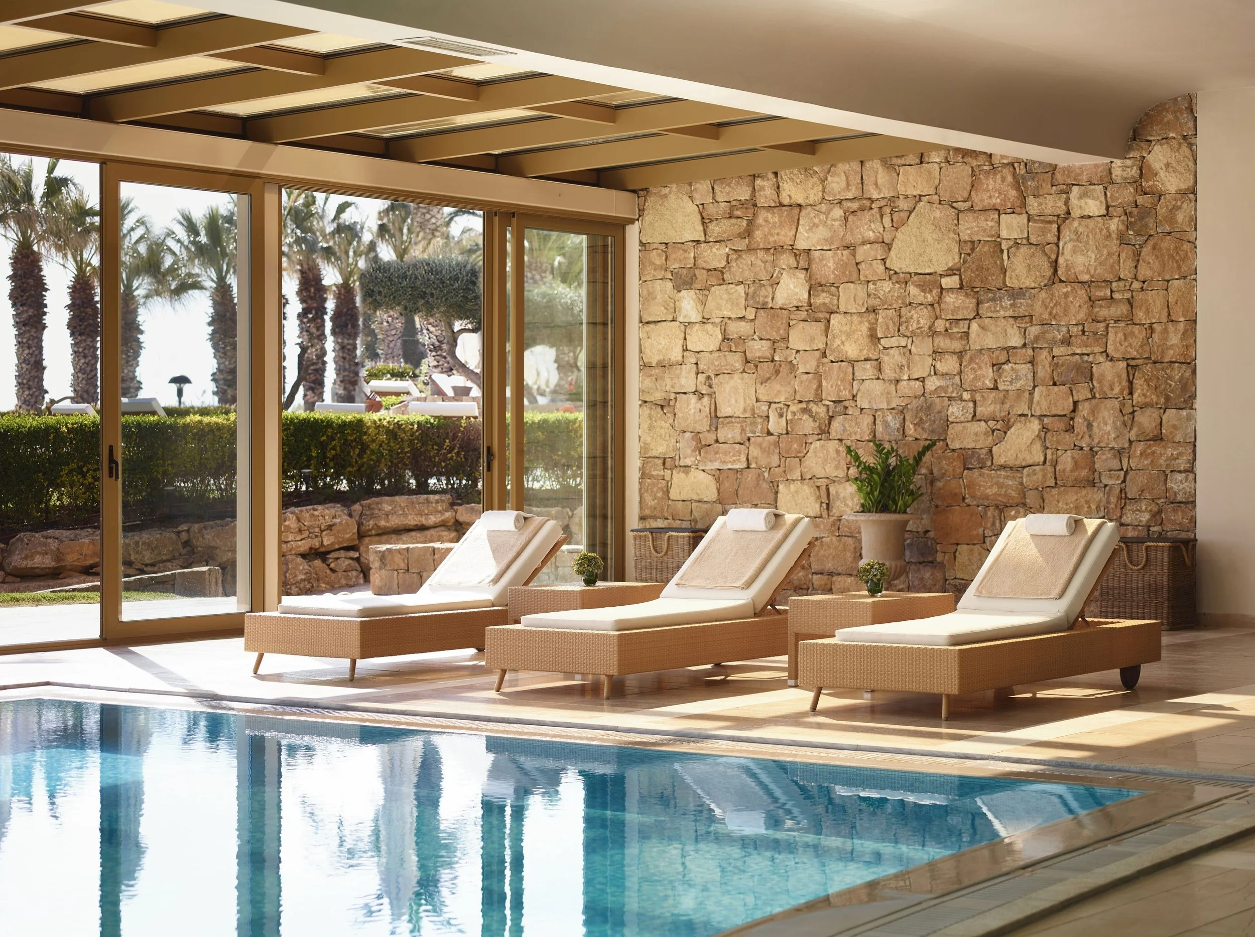 Indoor swimming pool area with lounge chairs, large glass sliding doors, a stone accent wall, and trees outside.
