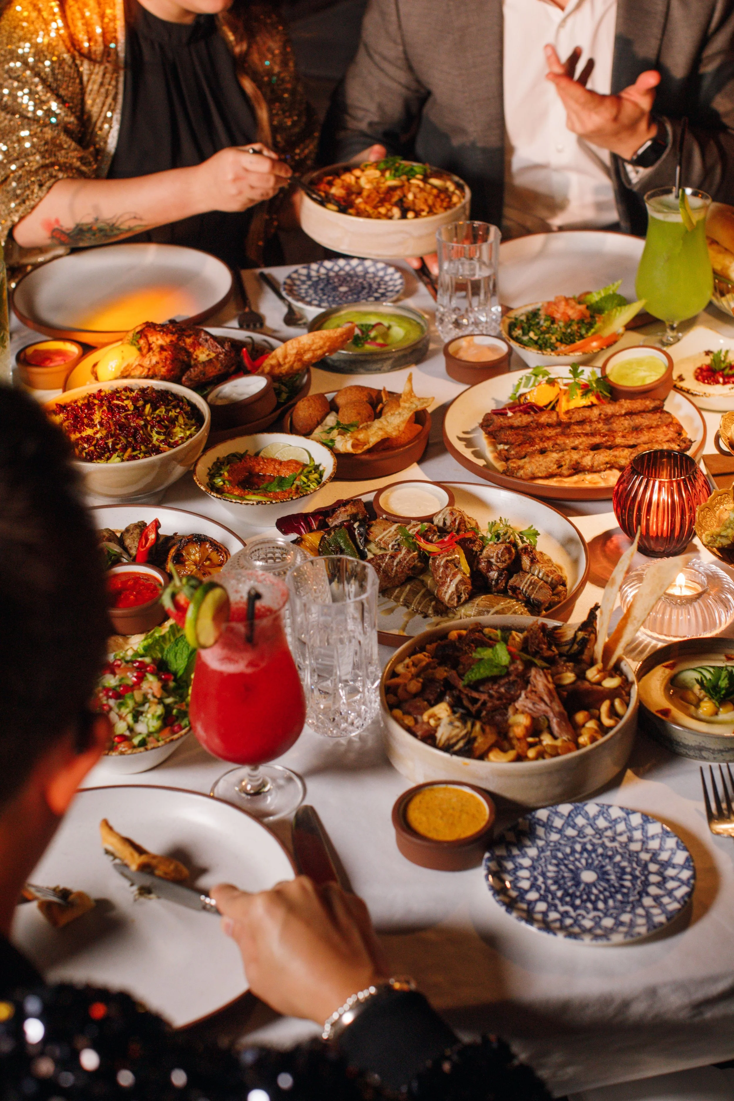 A table set with various dishes including grilled meats, salads, breads, and colorful drinks. People are serving and enjoying the meal, with some holding bowls and utensils.