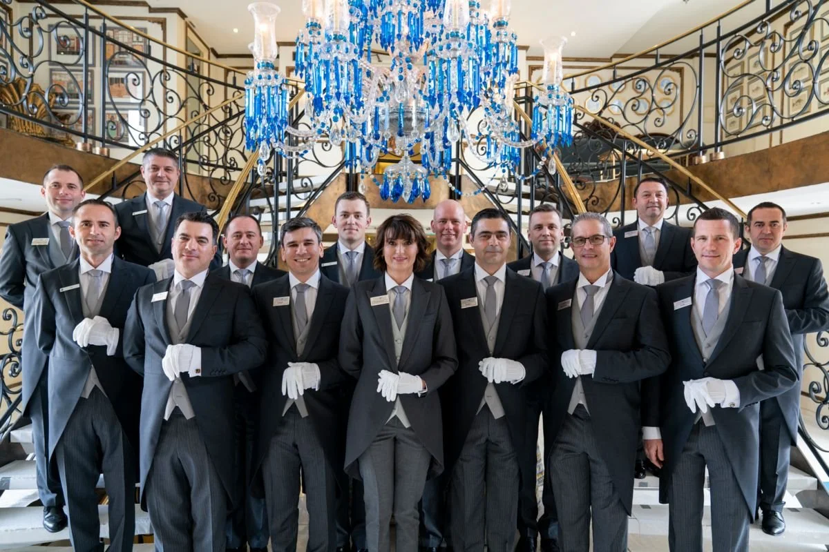 A group of sixteen hotel staff members, including a woman and fifteen men, dressed in formal uniforms with white gloves, standing on a staircase underneath a large blue and crystal chandelier in an elegant hotel lobby.