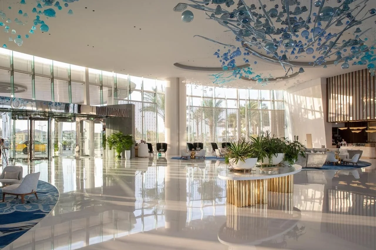 A spacious hotel lobby with large floor-to-ceiling windows, modern furniture, and decorative blue glass art on the ceiling.