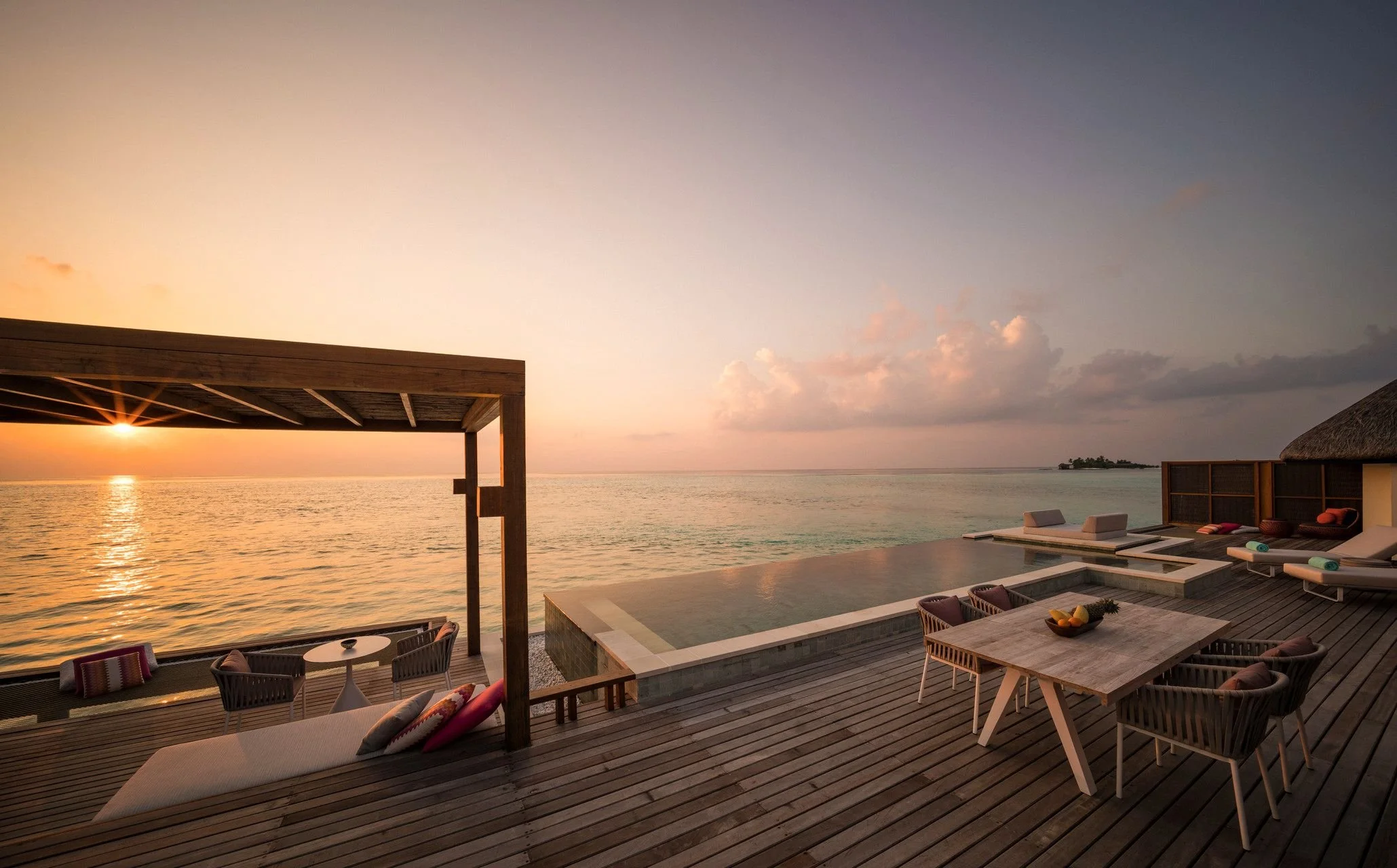 An outdoor lounging area with a view of the ocean at sunset. There are chairs, a table with fruit, and a pool with infinity edge.