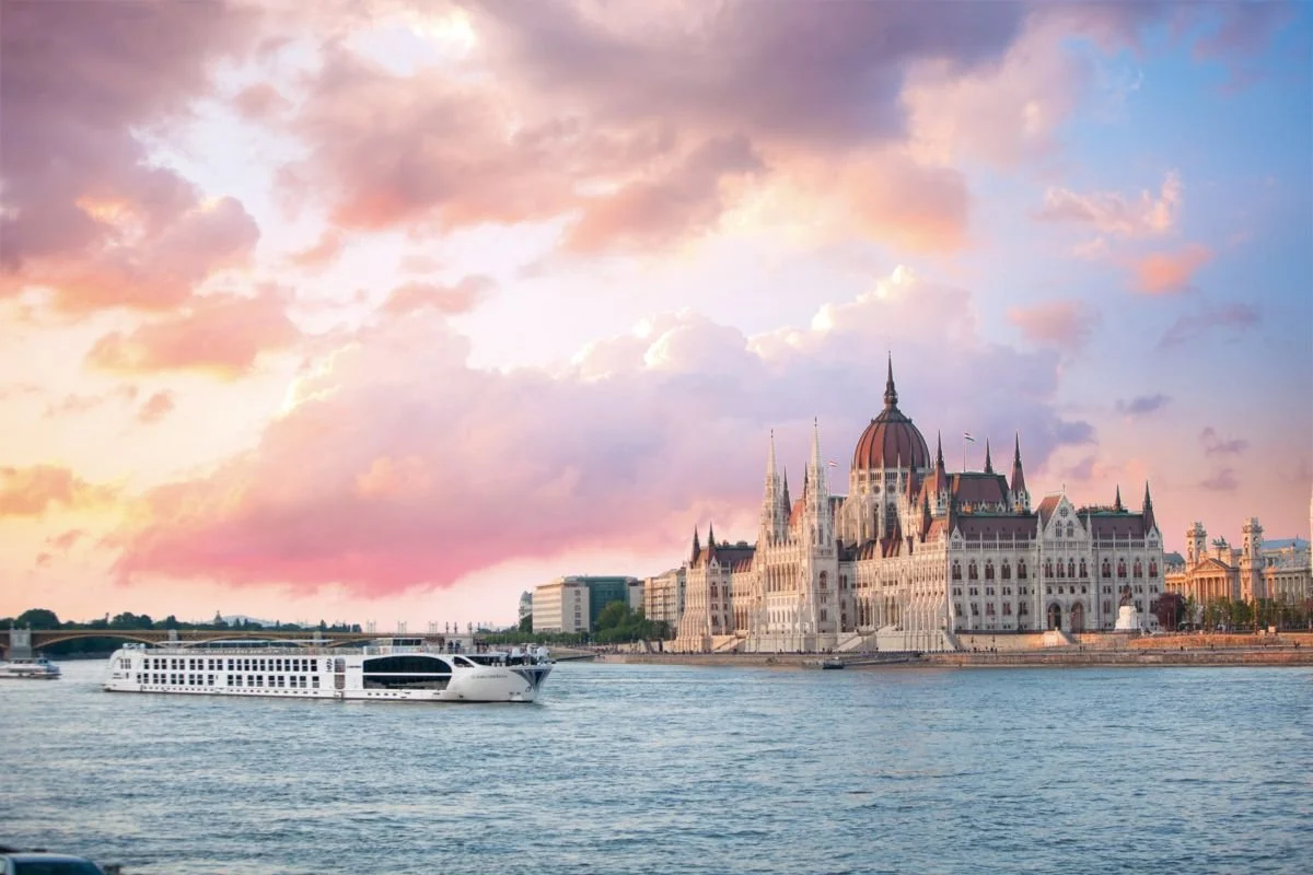 A cityscape featuring the Hungarian Parliament Building on the bank of the Danube River during sunset, with a white cruise boat in the water and pink and purple clouds in the sky.