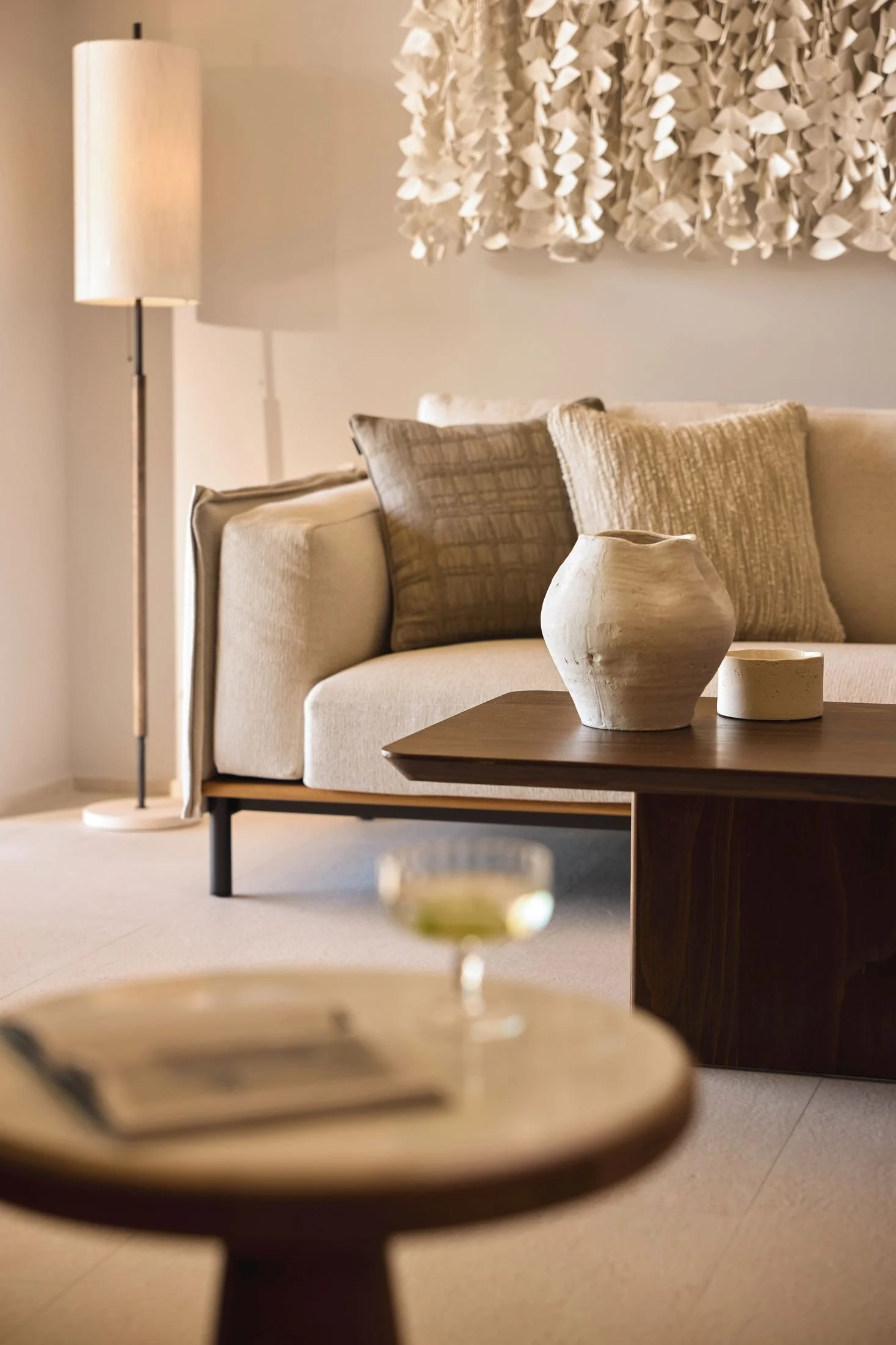 Living room with a beige sofa, a wooden coffee table with decorative white vases, beige pillows, a tall floor lamp, a wall hanging, and a side table with a drink and magazines.
