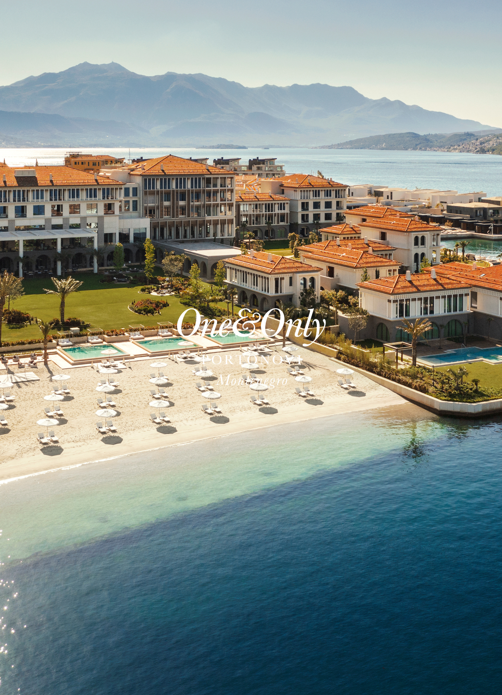 Aerial view of a beach resort with white sandy beach, turquoise water, and resort buildings with orange tiled roofs, with mountains and a lake in the background.