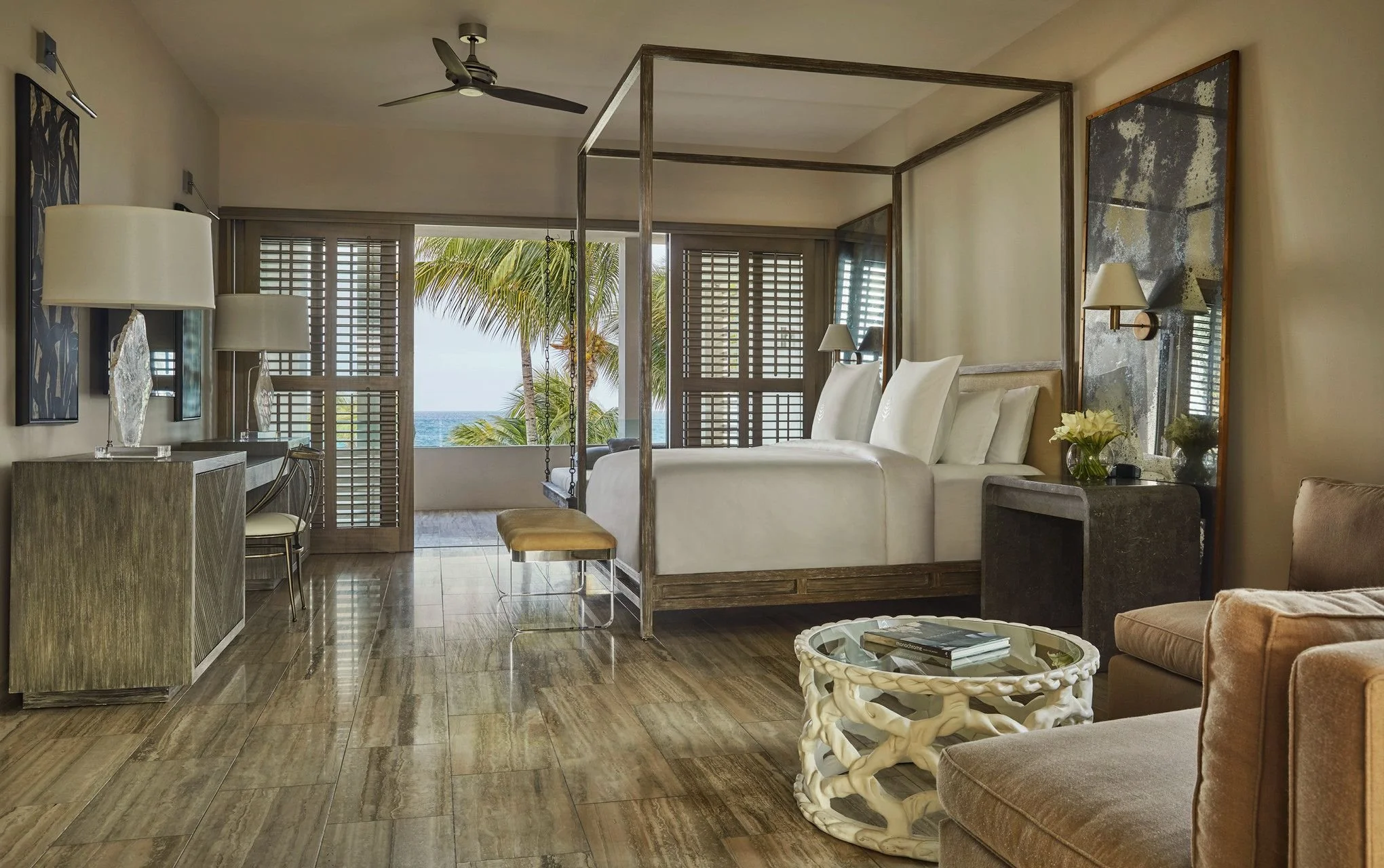 A tropical hotel room with a large window showing palm trees and the ocean, a bed with white linens, a wooden canopy frame, a lounge chair, a glass-top side table, and modern decor including artwork and lamps.