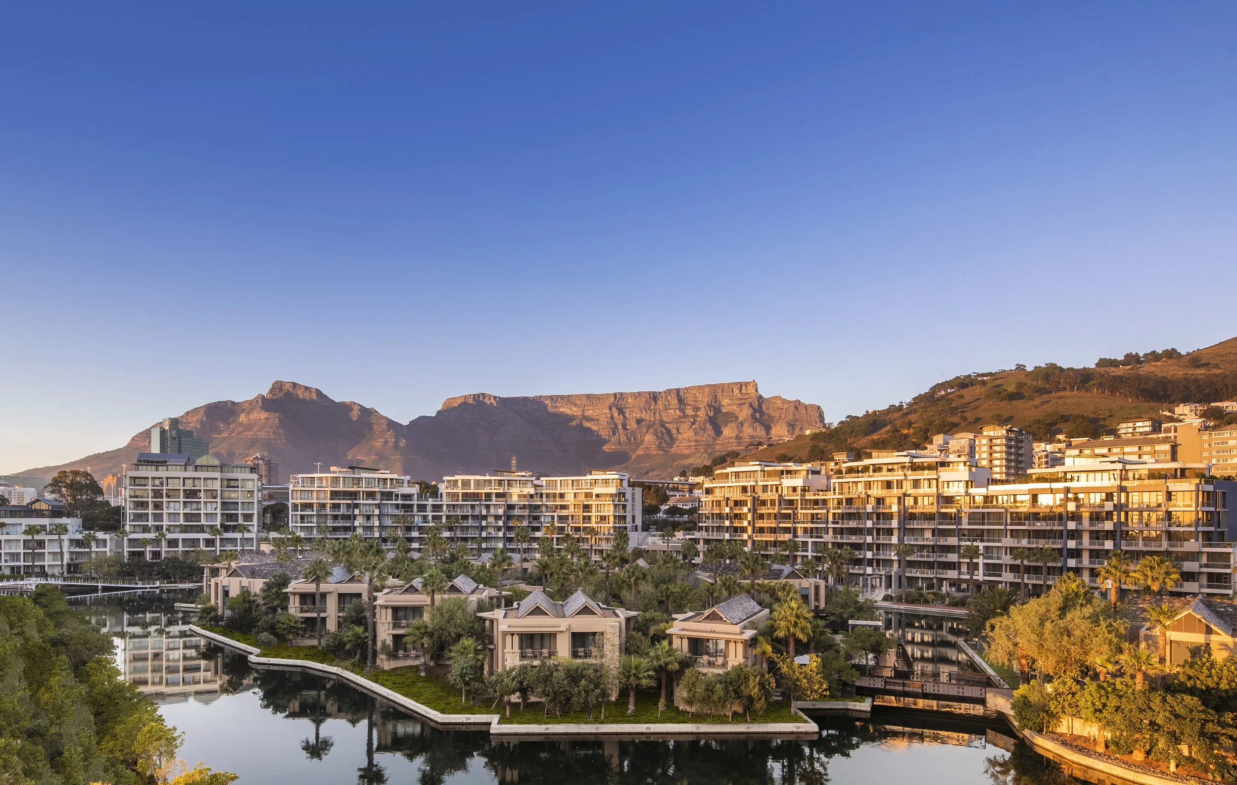 Cityscape with modern residential buildings, a water canal, trees, and a mountain range in the background under a clear blue sky at sunset.