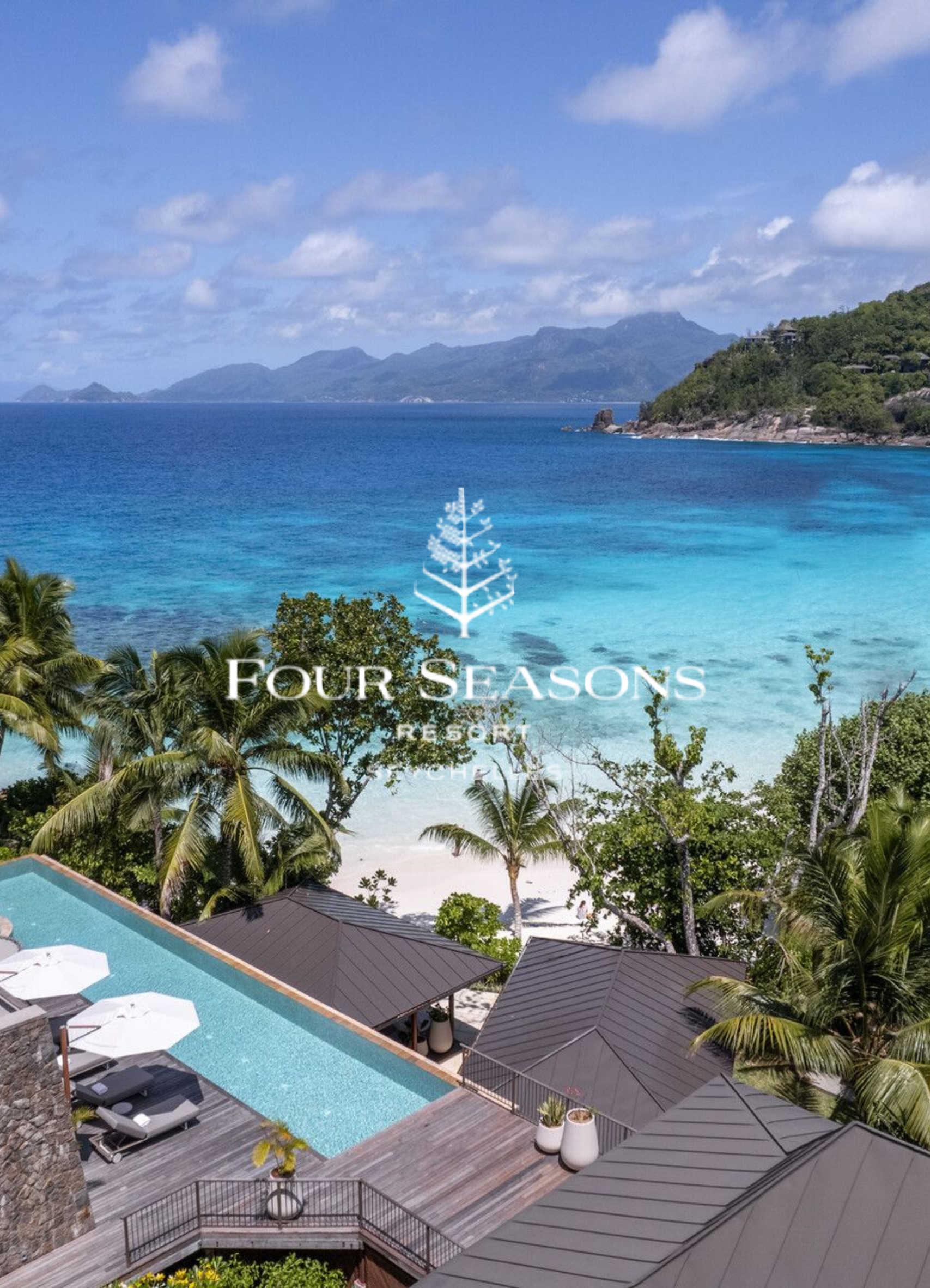 A coastal resort with a swimming pool, lounge chairs, and umbrellas in the foreground, palm trees and buildings around it, and a large body of water with mountains in the background under a partly cloudy sky.