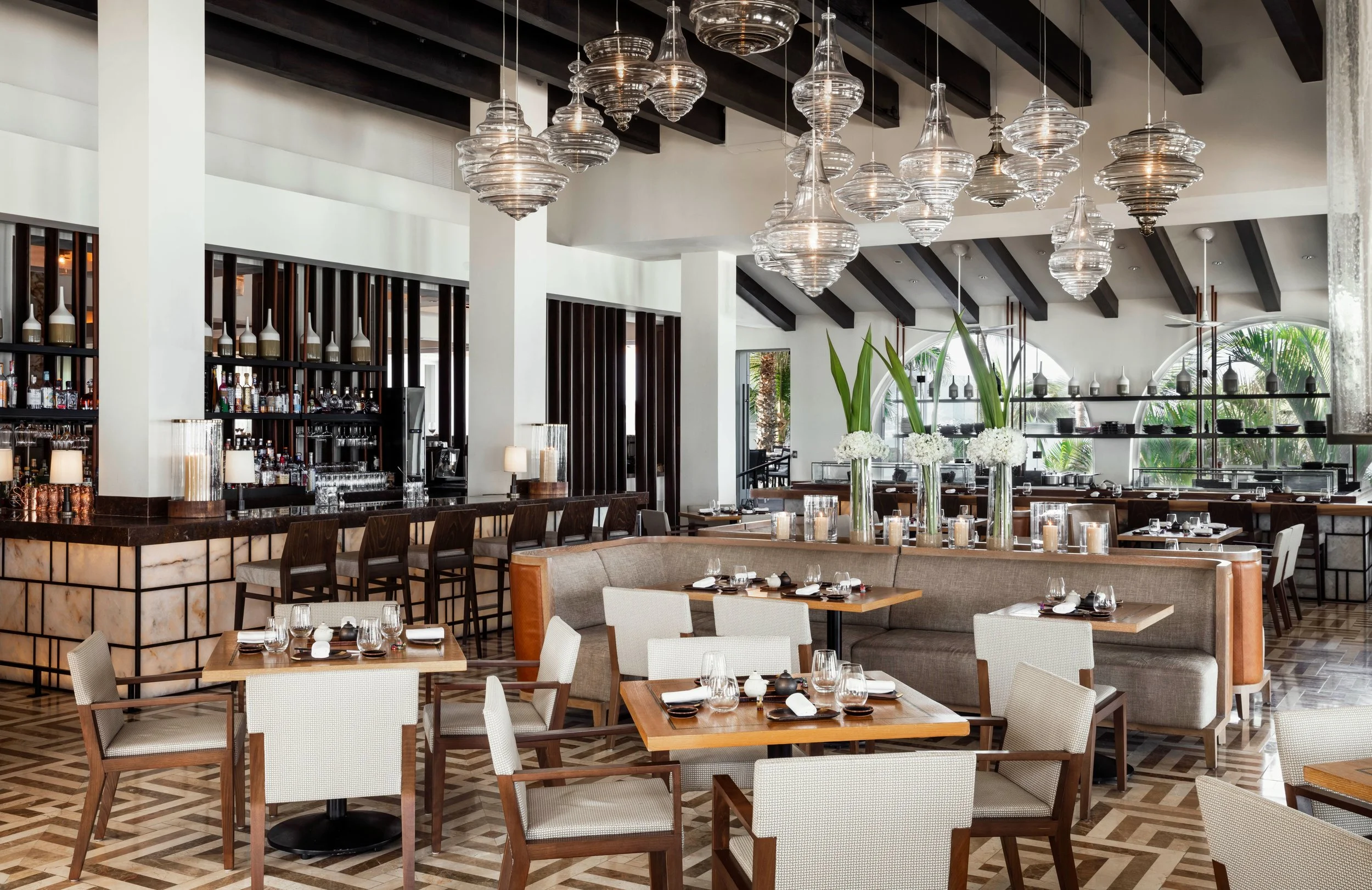Interior of a modern upscale restaurant with a bar area, dark wood accents, white walls, glass pendant lights, and neatly set tables with white and beige chairs.