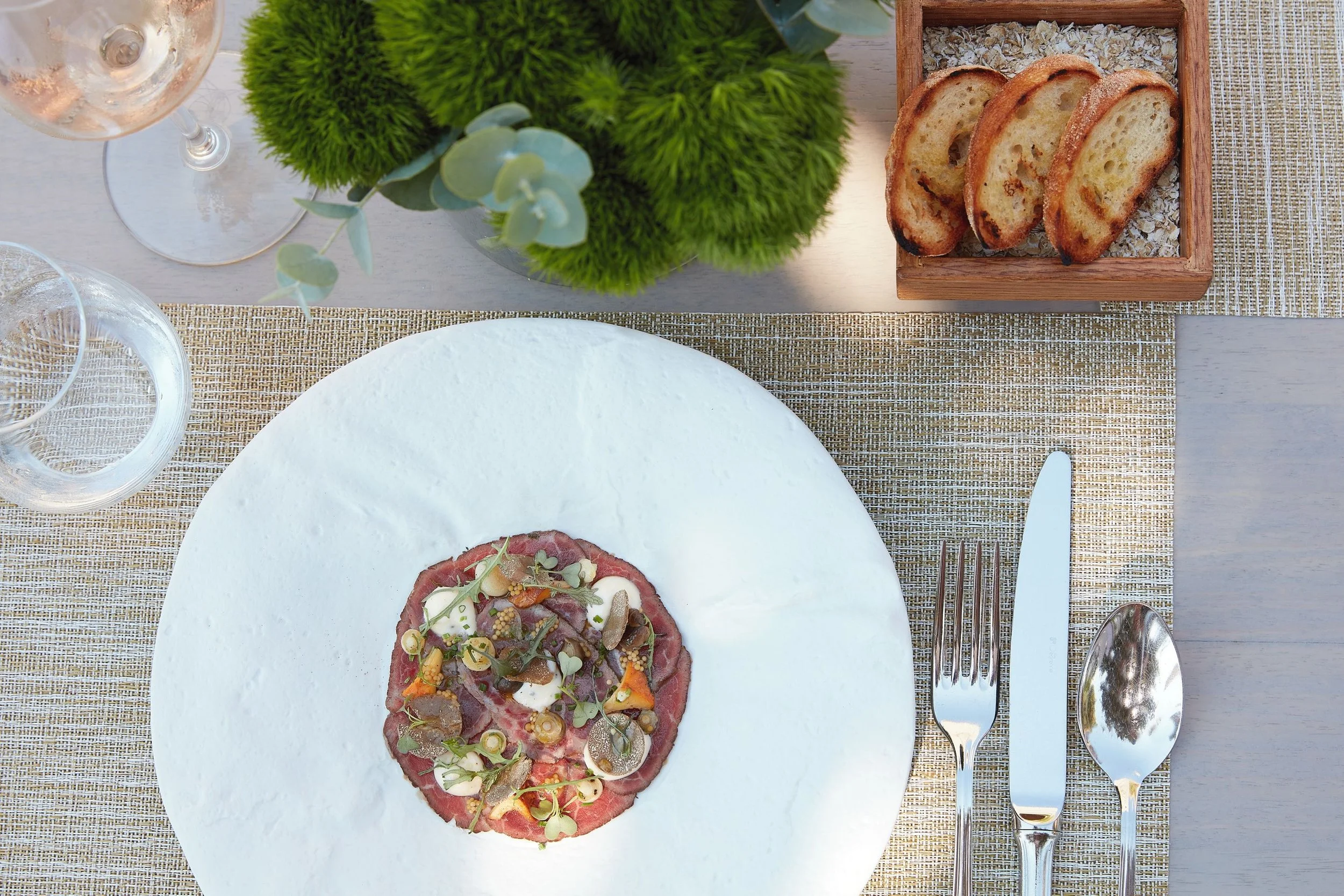 A plate of steak tartare garnished with microgreens, chopped pickles, and mustard seeds, placed on a white plate on a woven placemat. Above the plate, there is a glass of white wine, a green floral arrangement, and a wooden box with toasted baguette slices. Silverware is set next to the plate.