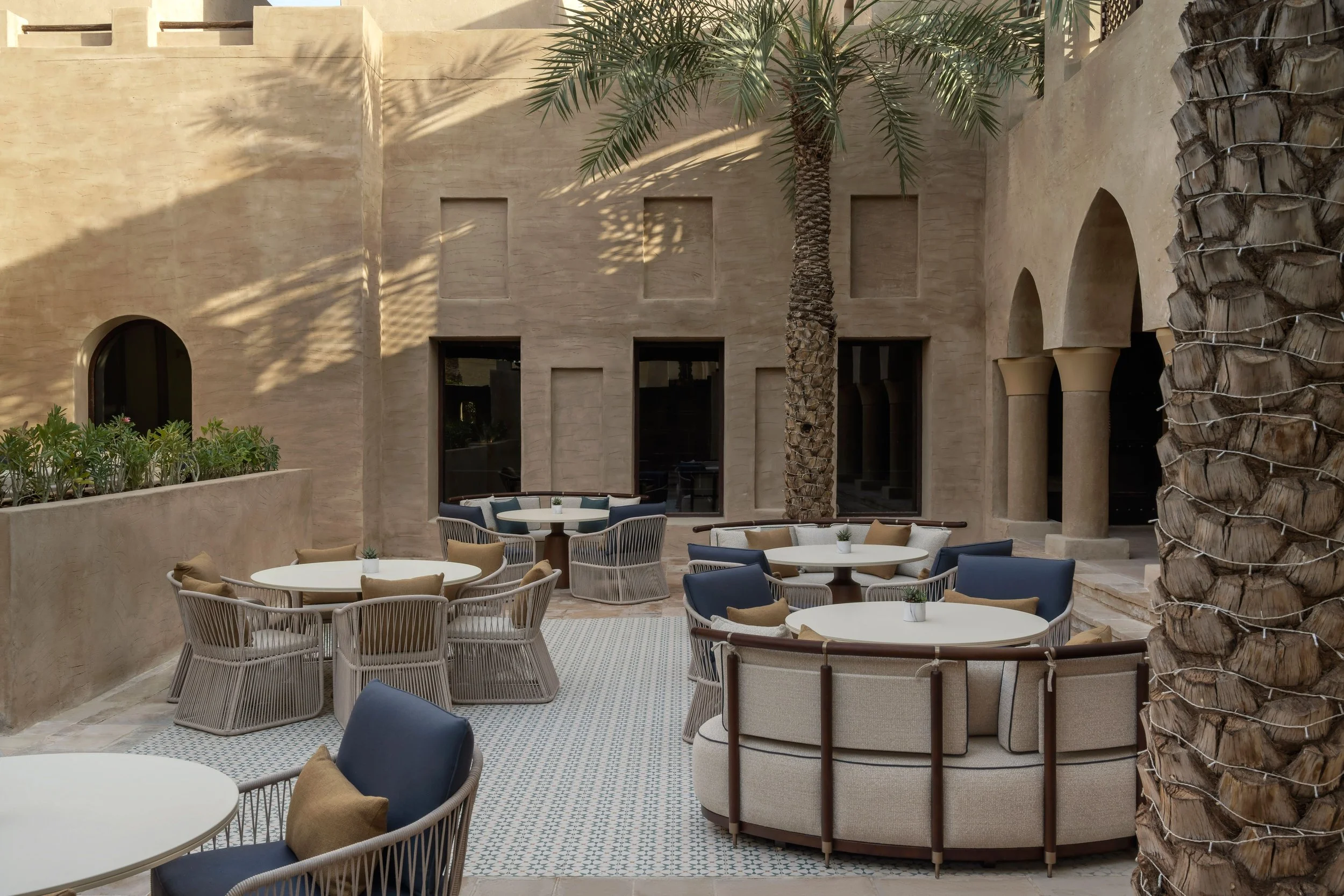 An outdoor patio area with tables and chairs, a large palm tree, and beige stone walls with arched windows and doorways.