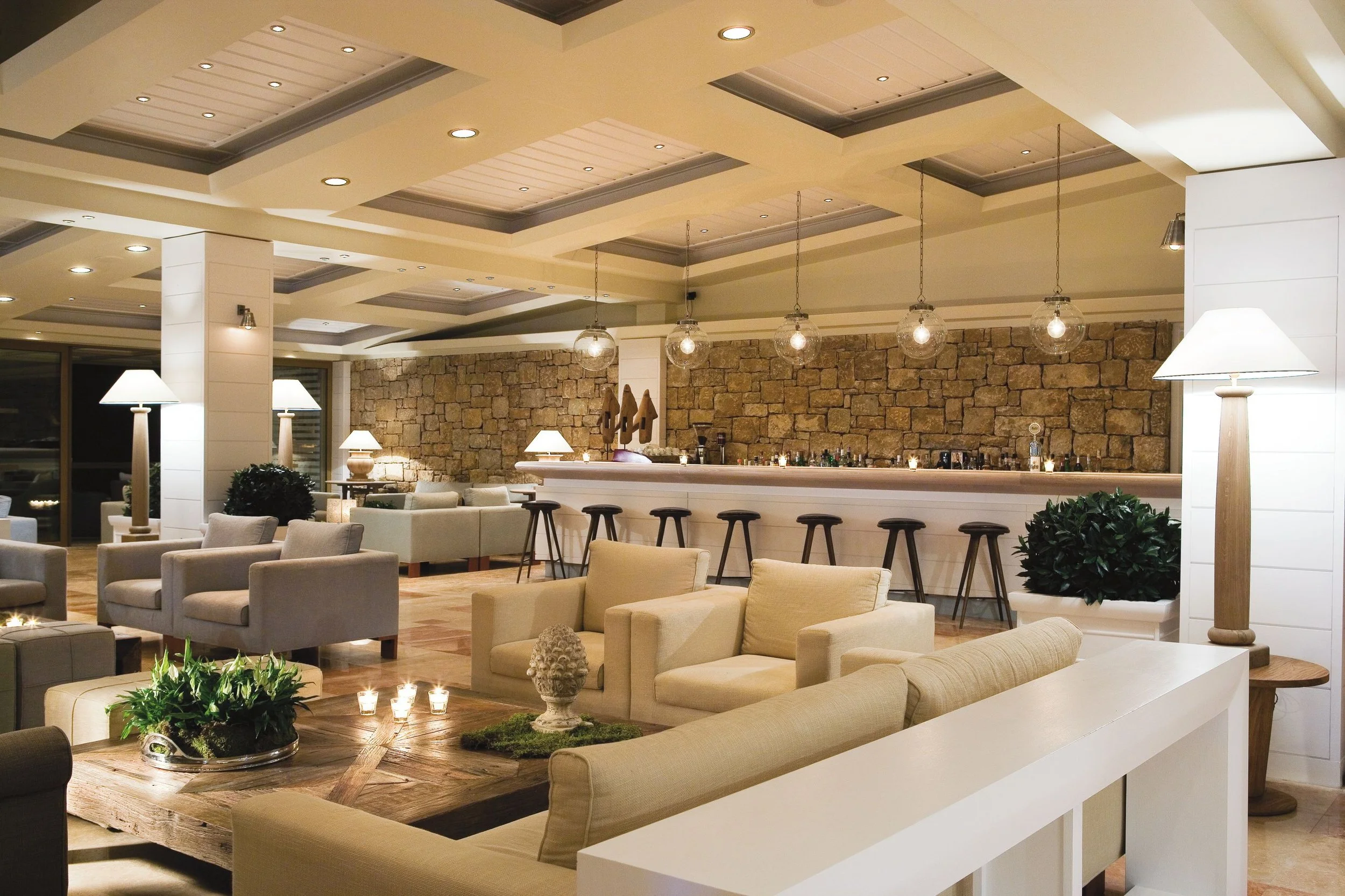 Interior of a modern lounge area with beige sofas, wooden coffee table, potted plants, multiple lamps, hanging globe lights, a stone accent wall, and a bar with barstools.