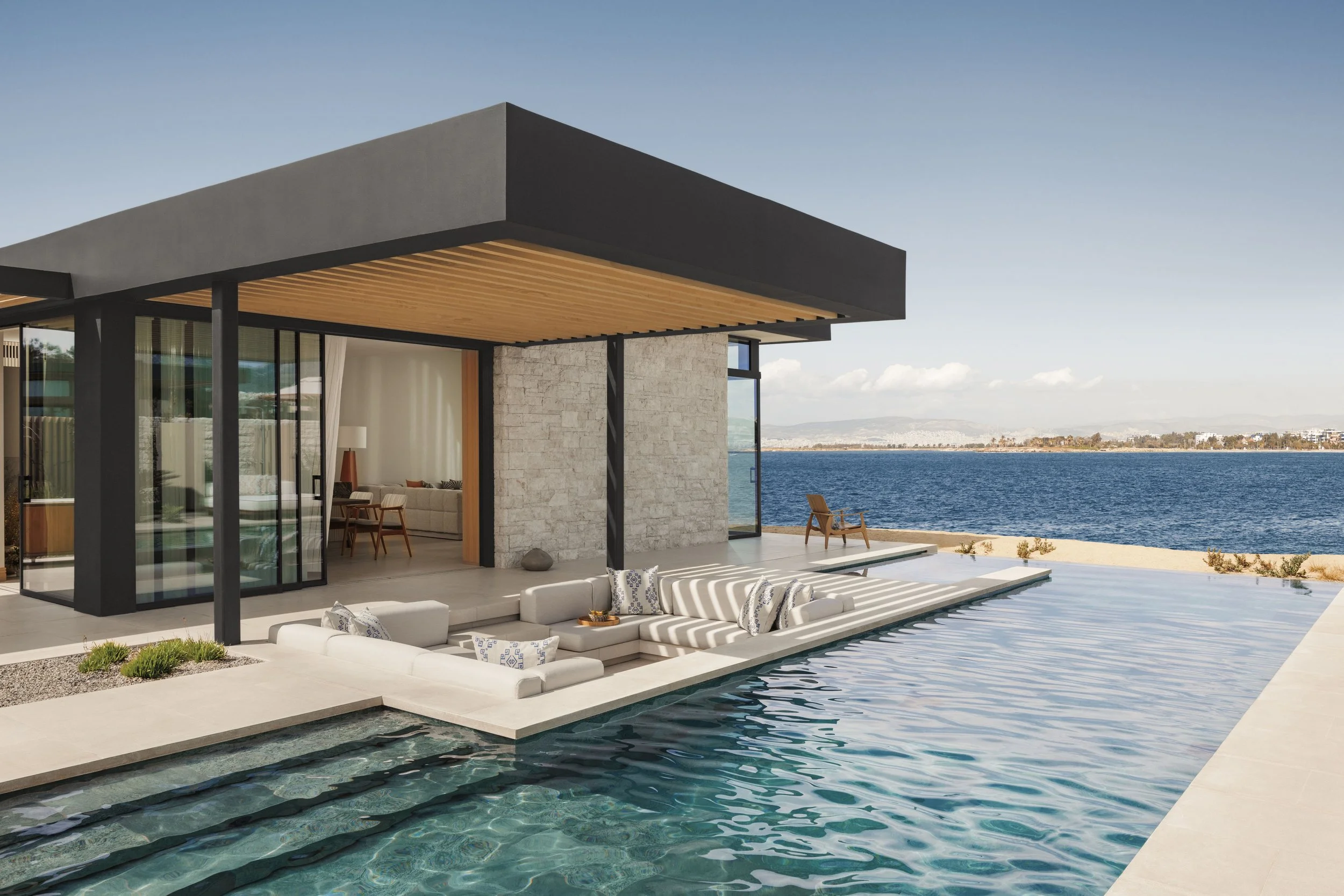 Modern house with large glass windows and sliding doors by a lake, with an outdoor seating area and pool.