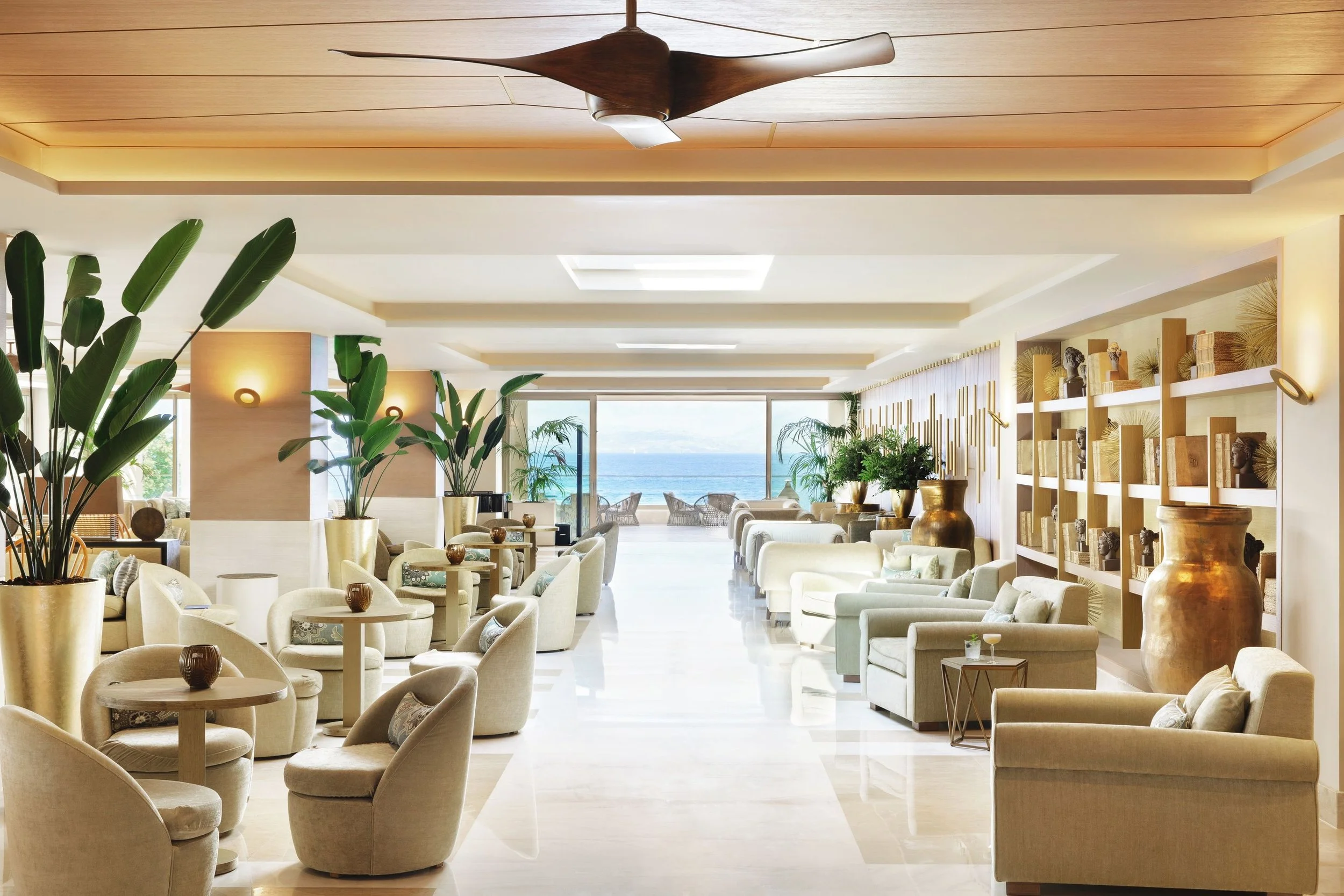 Luxurious hotel lobby with beige armchairs and sofas, potted plants, and decorative items. Large windows reveal a view of the ocean in the background. The ceiling features wood and white paneling with a modern ceiling fan.