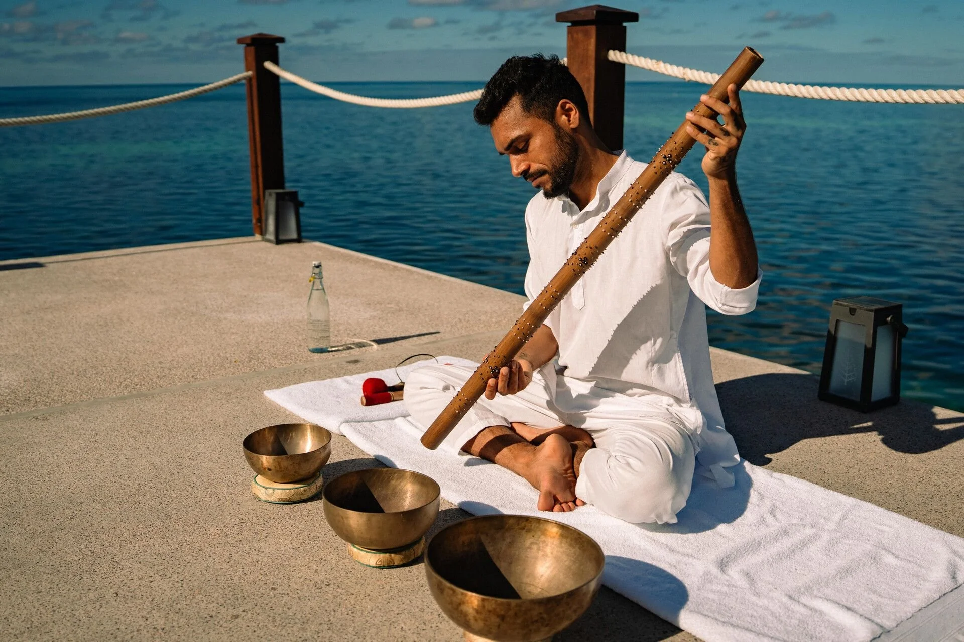 A man sitting cross-legged on a white towel by the ocean, playing a wooden flute with metal embellishments, surrounded by four brass bowls, with a bottle of water and a black lantern nearby.